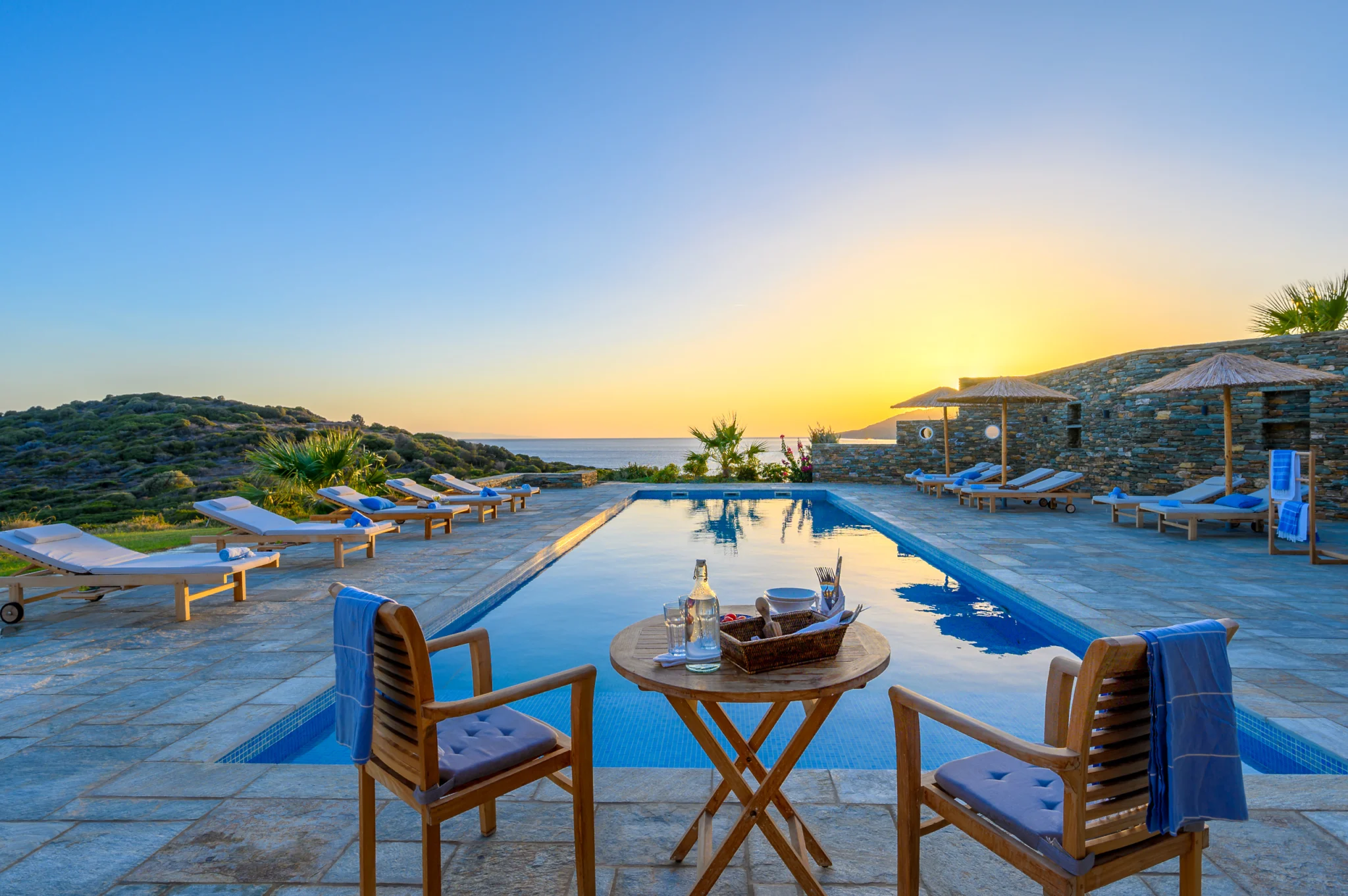 Villa Golden Beach — Evening poolside seating, Andros Poolside timber chairs and a table with drinks beside the reflective pool at sunset at Villa Golden Beach in Andros