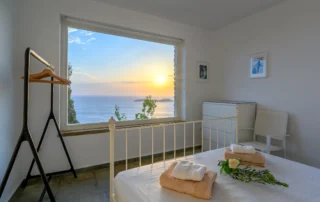 White bedroom with a large window framing sunset over the sea at Villa Hermes, Andros, Greece