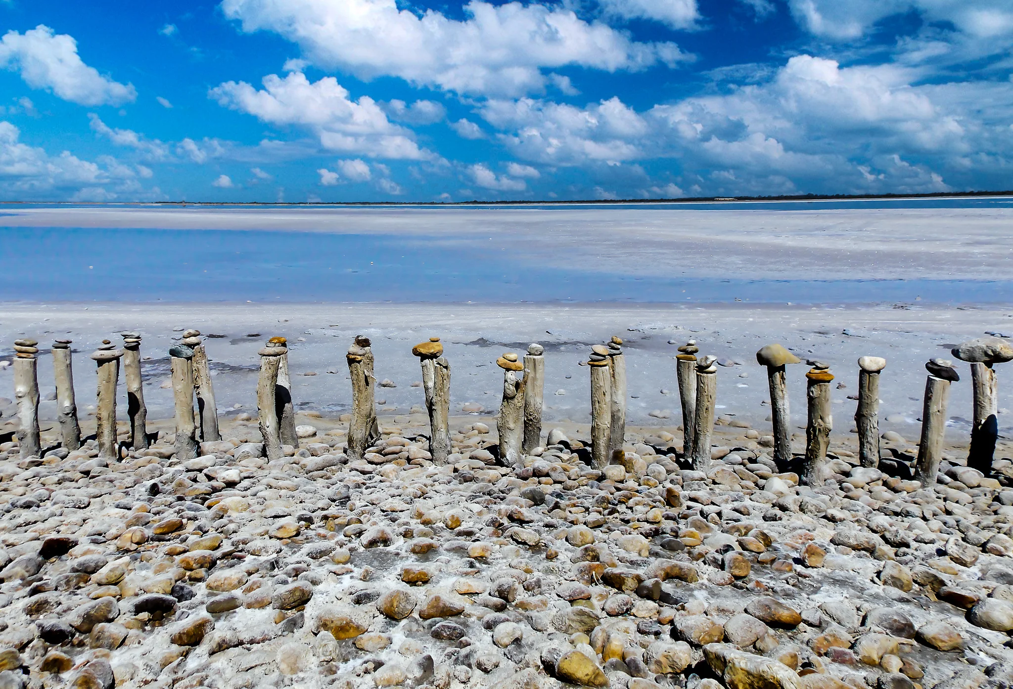 Wooden posts topped by balancing stones along a pale lakeshore