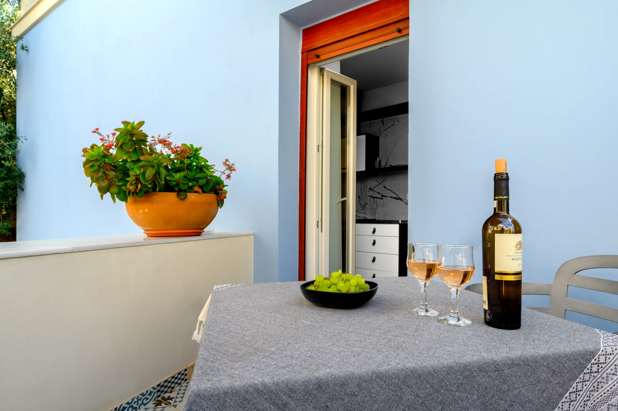 Outdoor dining table with wine and a flowering plant beside a blue wall at Blue Sky House, Pylos, Peloponnese, Greece.