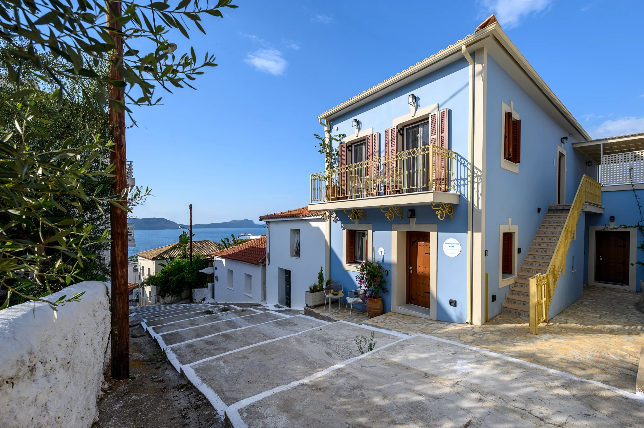 Blue house and paved forecourt framed by an olive tree at Blue Sky House, Pylos, Peloponnese, Greece.
