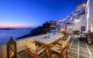 Marble-topped dining table, timber chairs and candle lanterns on a pebble-paved terrace overlooking the sea and illuminated Fira cliffs at blue hour at Boutique Hotel Fira in Santorini.