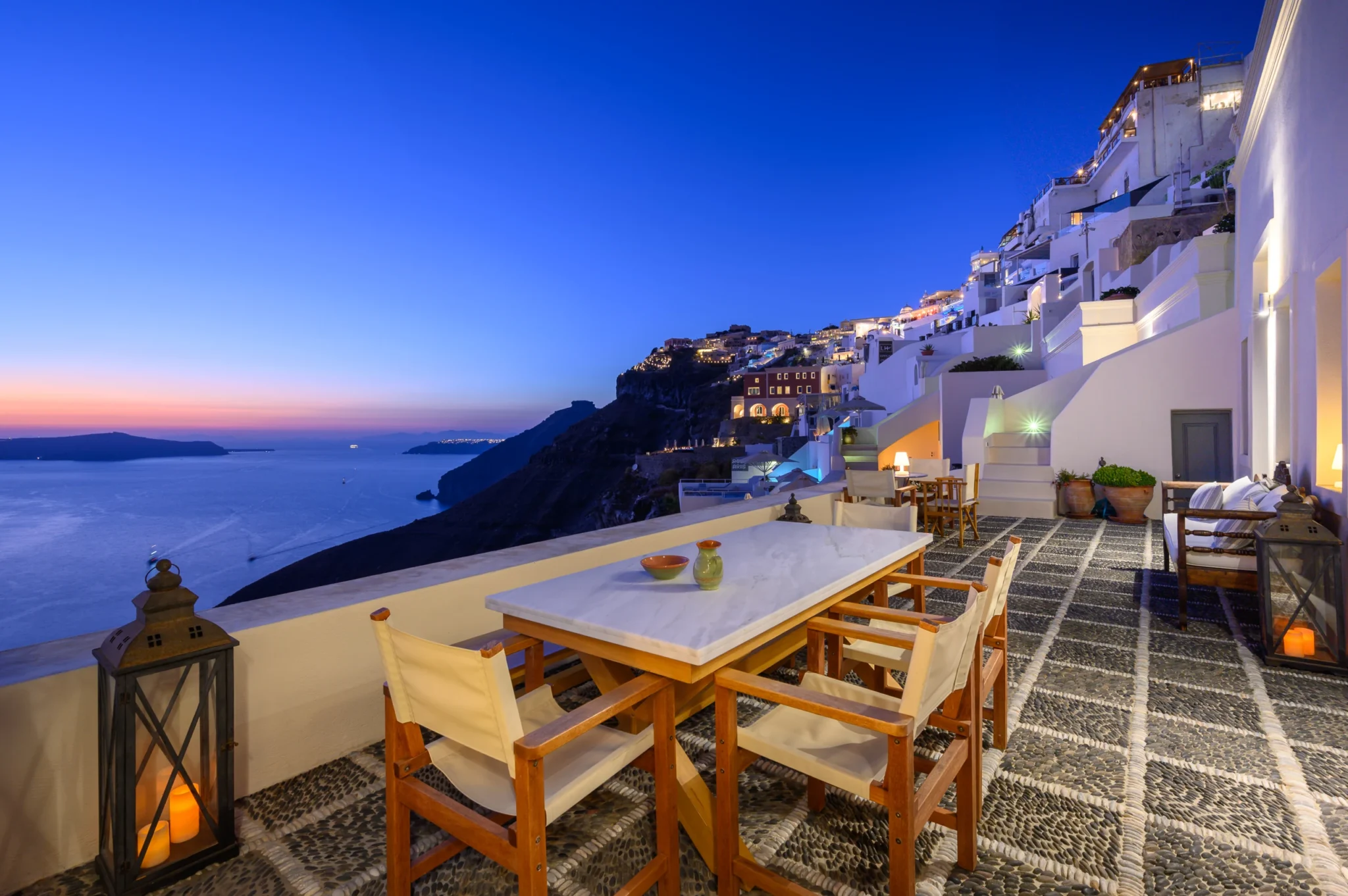 Marble-topped dining table, timber chairs and candle lanterns on a pebble-paved terrace overlooking the sea and illuminated Fira cliffs at blue hour at Boutique Hotel Fira in Santorini.
