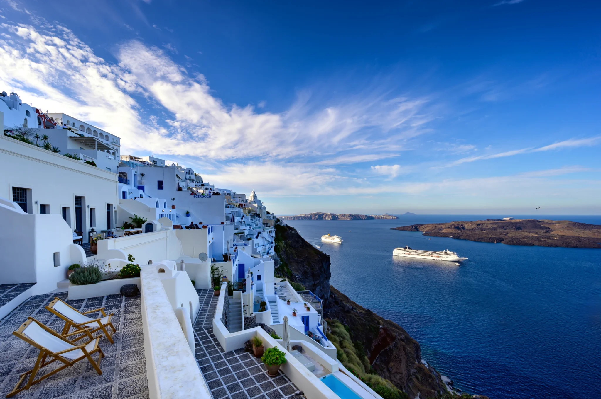 Deckchairs on a pebble-paved terrace overlooking white cliffside buildings and the caldera with cruise ships at Boutique Hotel Fira in Santorini.