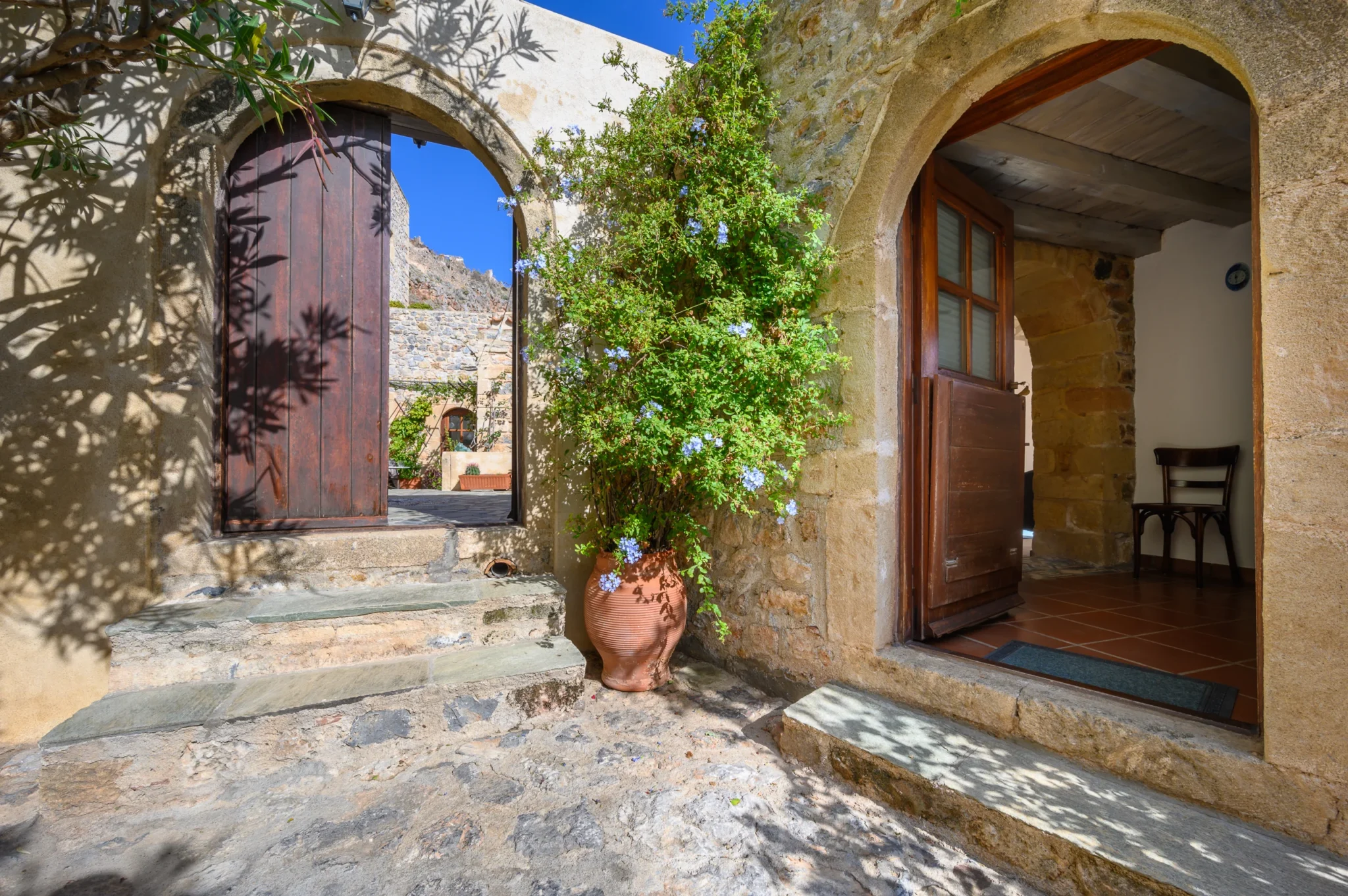 Open arched timber gate and residence doorway beside stone steps and a flowering climber in a terracotta pot at Castle Residence in Monemvasia.