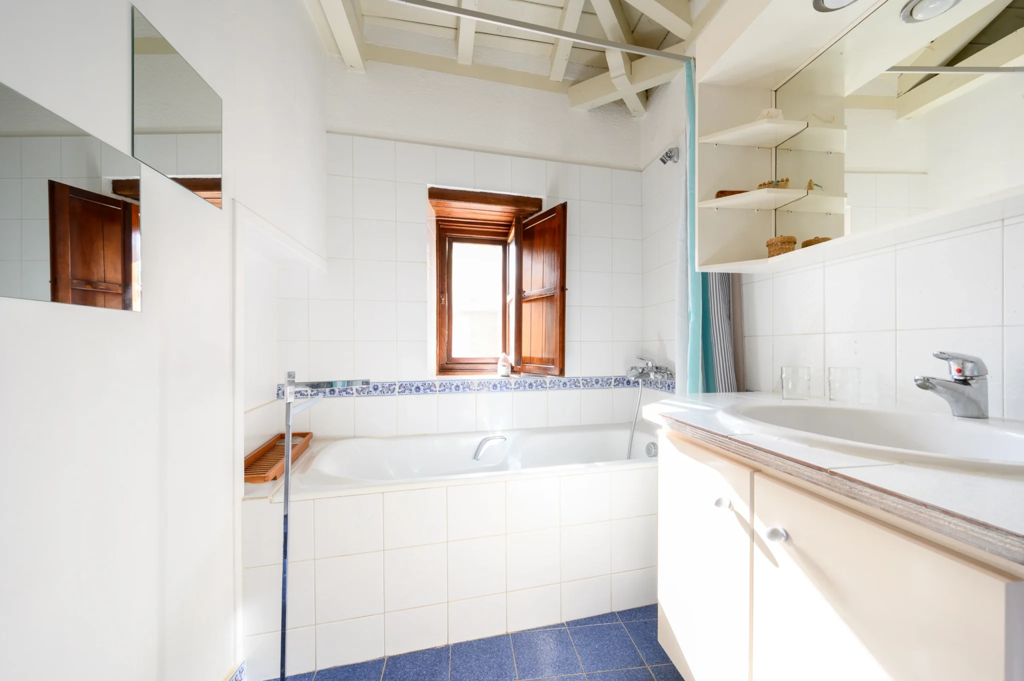 White-tiled bathroom with a bathtub, glass shower screen, timber window, white vanity and blue tile details at Castle Residence in Monemvasia.