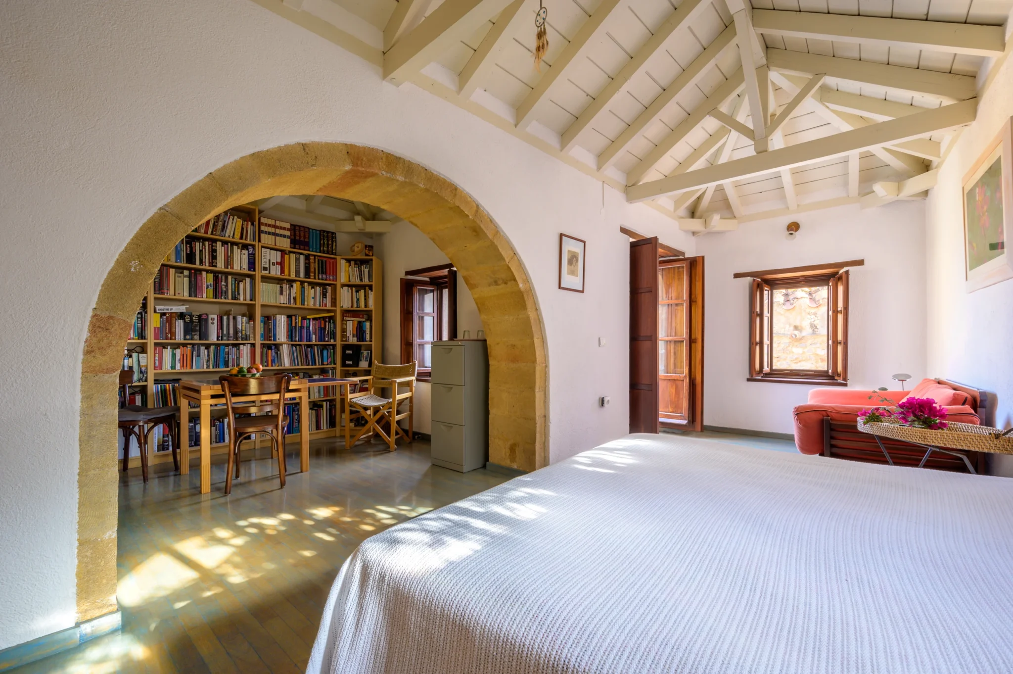 White bed and coral sofa beneath an exposed timber roof, with a stone arch framing the adjoining library and table at Castle Residence in Monemvasia.