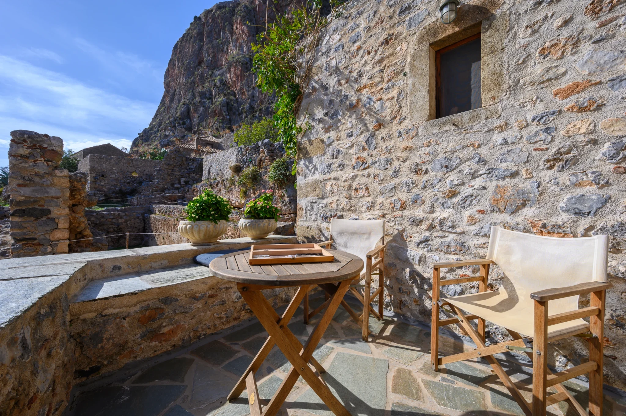 Round timber table with a backgammon board and canvas chairs beside a stone facade, with Monemvasia rock rising beyond at Castle Residence in Monemvasia.