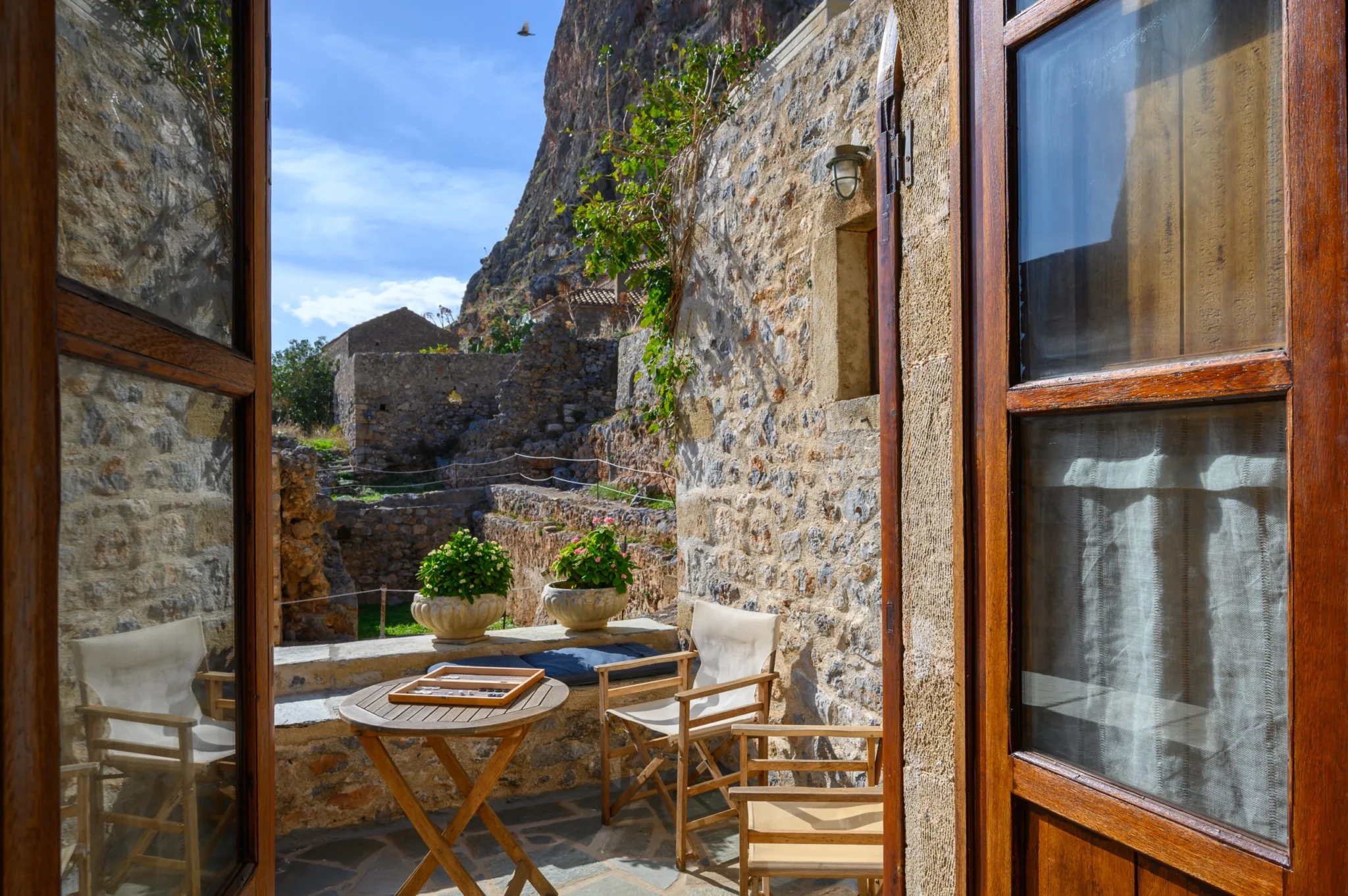 Open timber doors framing a stone courtyard with a round table, backgammon board, canvas chairs and potted flowers beneath Monemvasia rock at Castle Residence in Monemvasia.