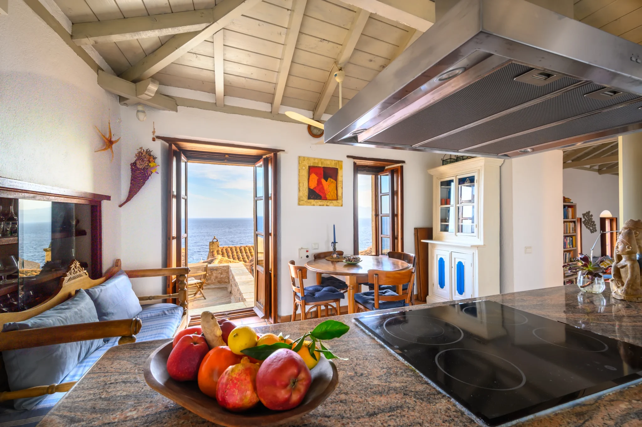 Kitchen island with a stone worktop and fruit bowl facing a timber dining table, white dresser and open sea view balcony doorway at Castle Residence in Monemvasia.