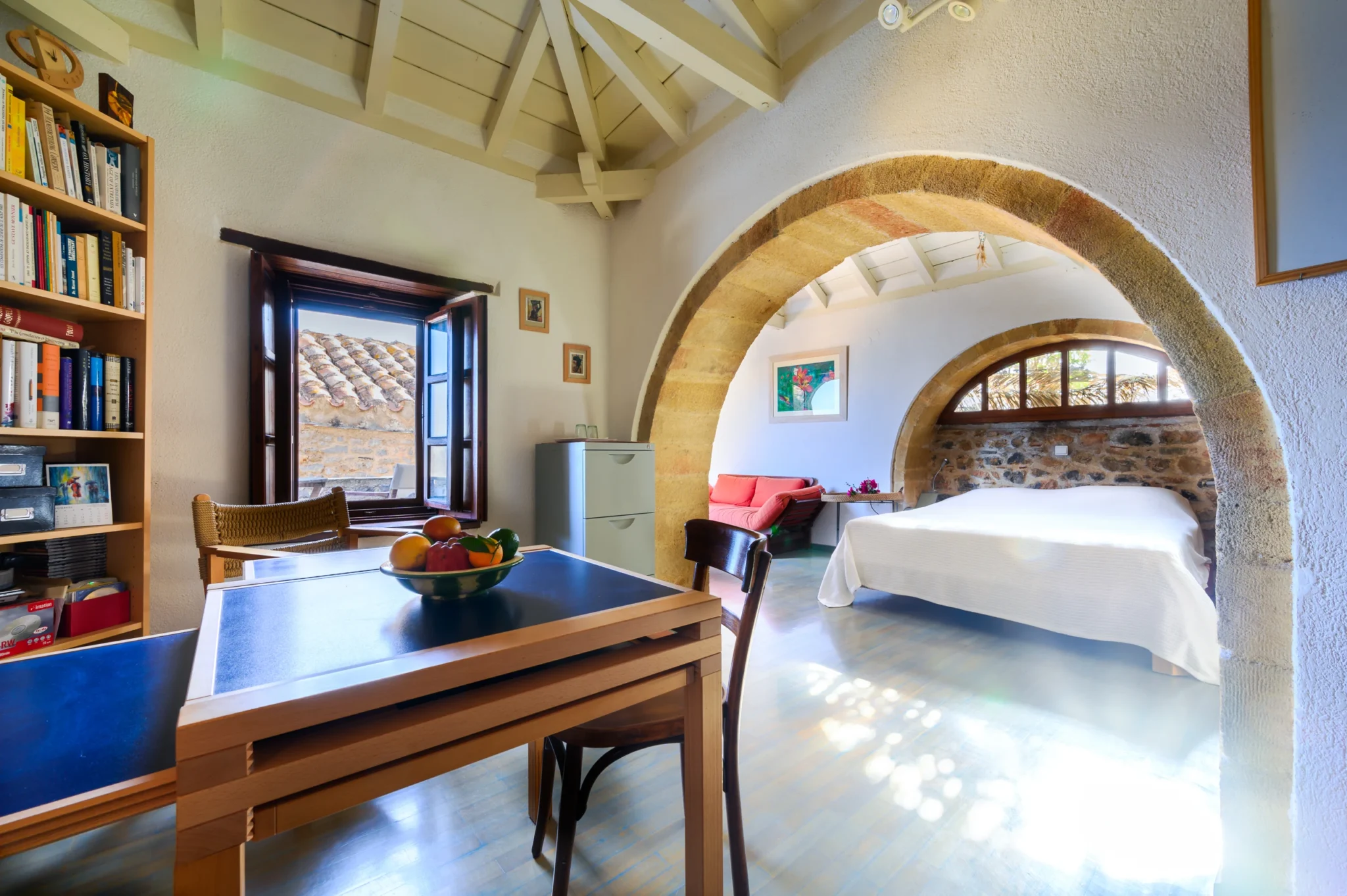 Timber table and bookshelves beside a broad stone arch opening to a bedroom with a white bed and curved window at Castle Residence in Monemvasia.