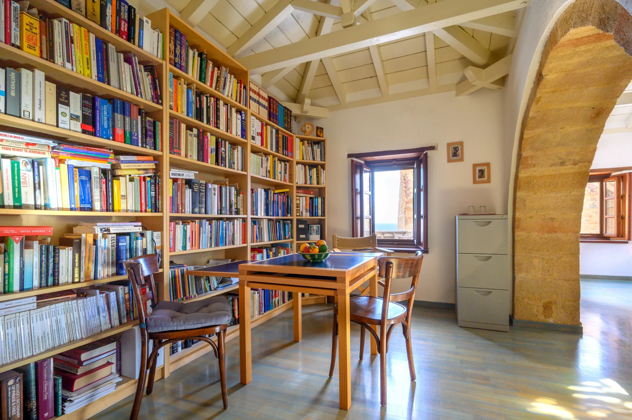 Library with wall-length bookshelves, a timber table and chairs, pale roof beams and a broad stone arch at Castle Residence in Monemvasia.