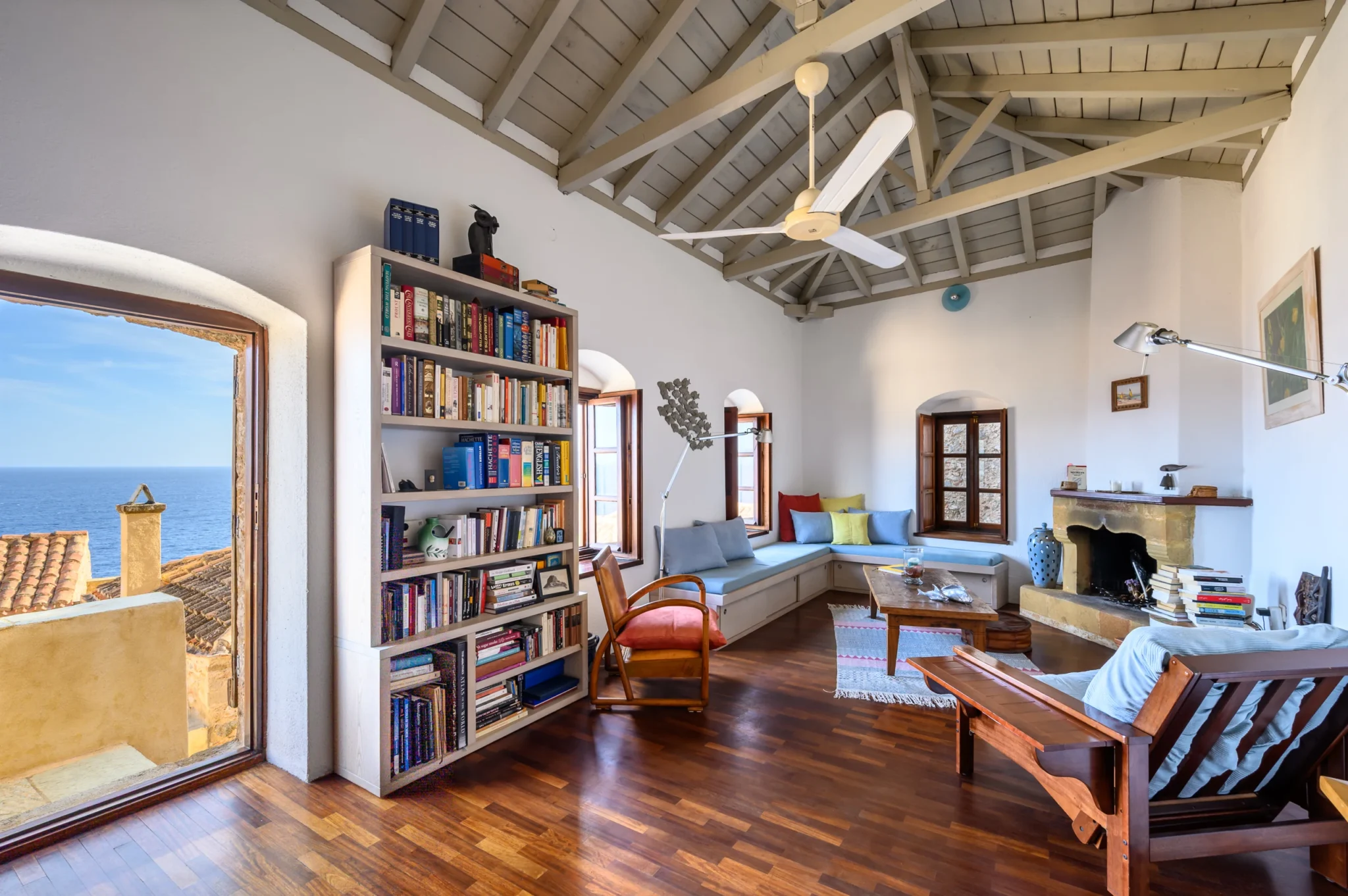 Living room with a bookcase, blue corner seating, stone fireplace and exposed timber roof beside an open balcony doorway overlooking the sea at Castle Residence in Monemvasia.