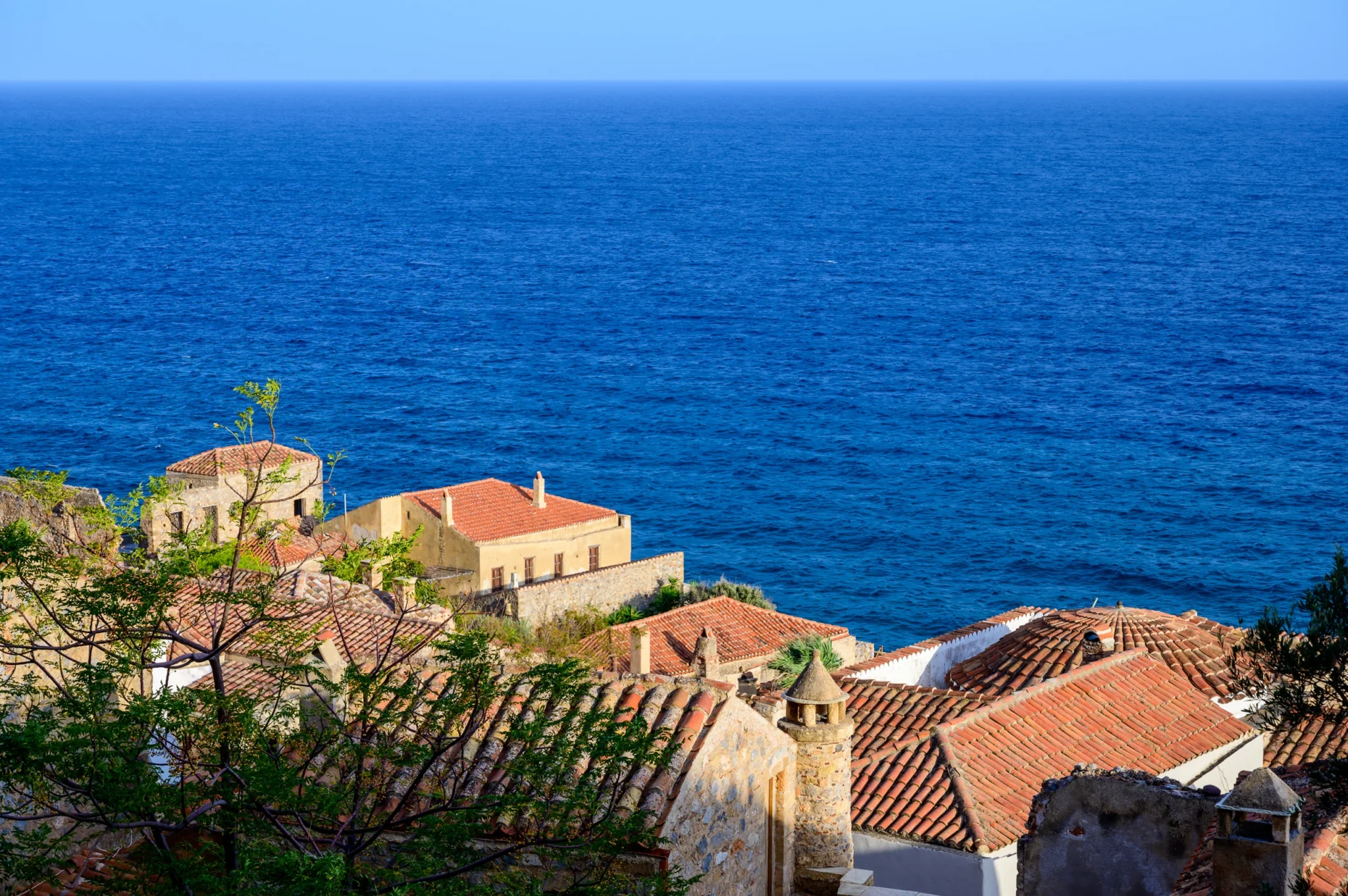Terracotta rooftops and stone buildings in Monemvasia beneath an expansive blue sea view, with tree branches in the foreground at Castle Residence in Monemvasia.