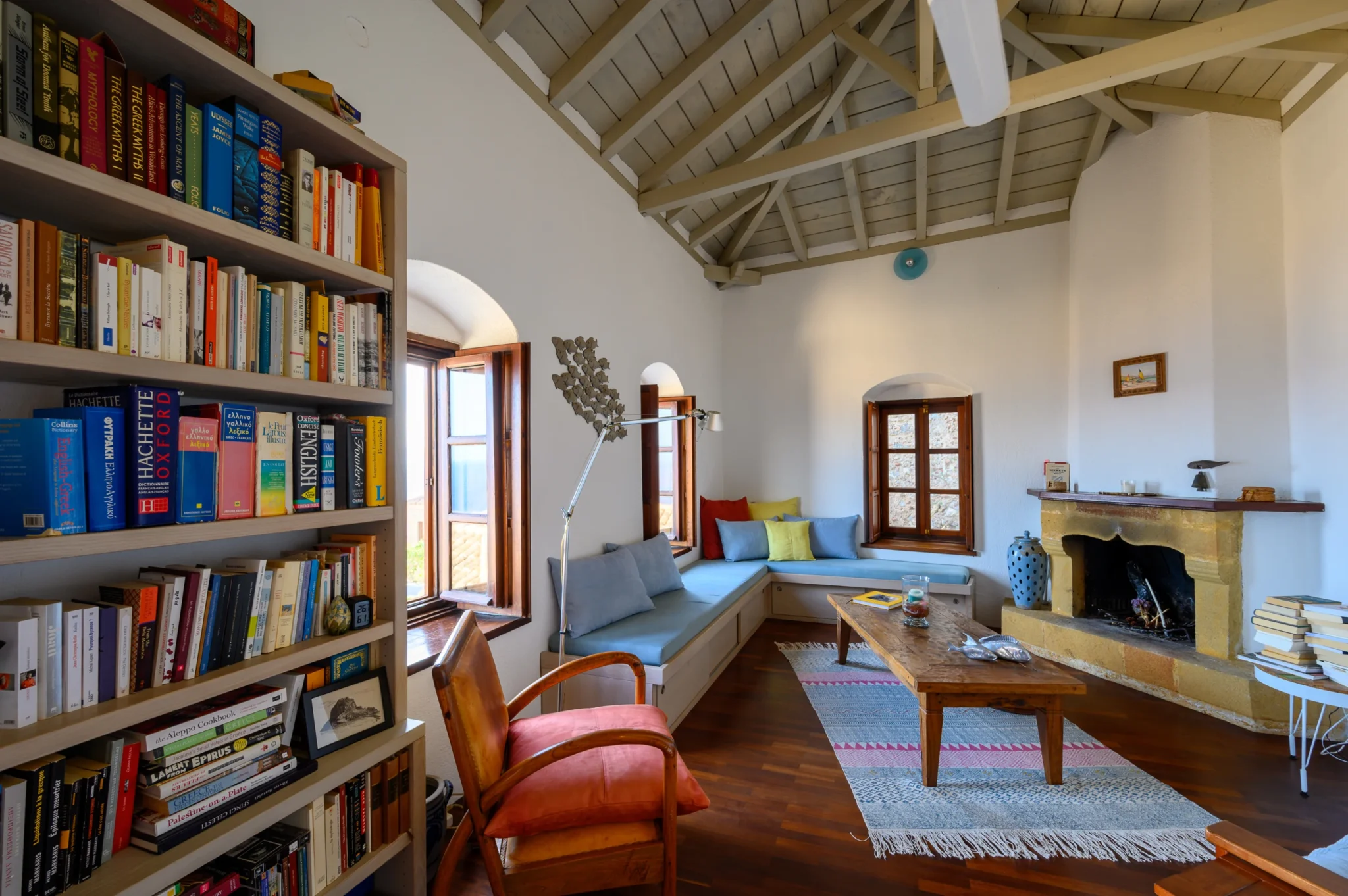 Book-lined living room with pale blue built-in seating, colourful cushions, a timber coffee table and a stone fireplace at Castle Residence in Monemvasia.