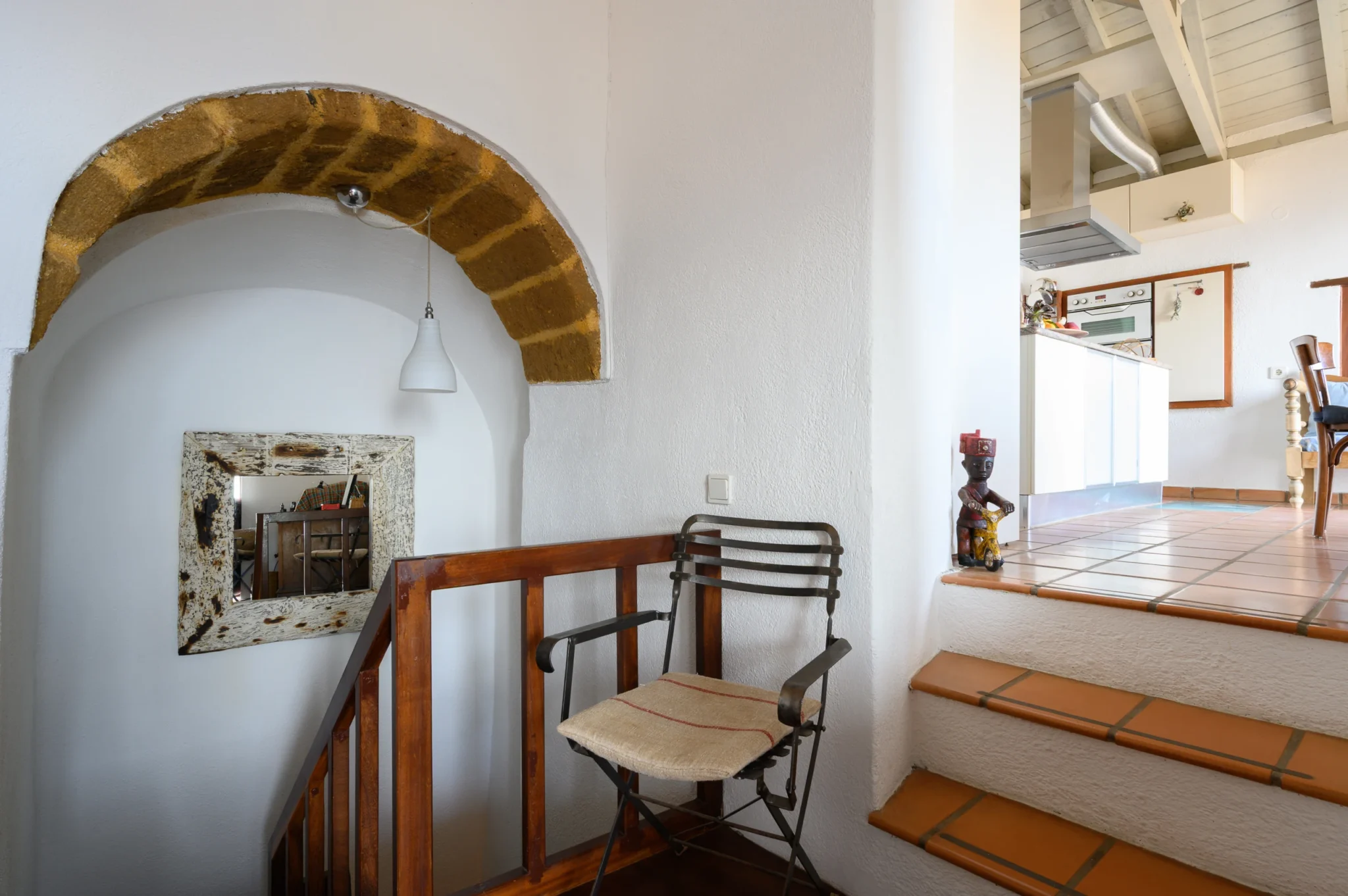 Stair landing with a stone arch, framed mirror and timber balustrade beside terracotta steps leading to the kitchen at Castle Residence in Monemvasia.