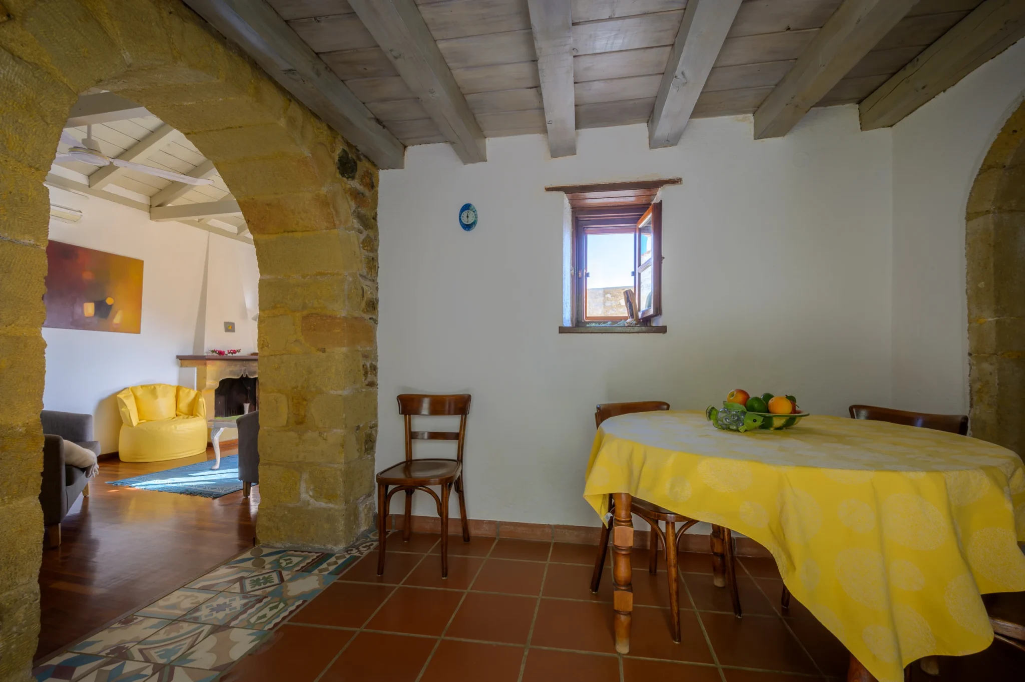 Dining table with a yellow tablecloth beneath timber ceiling beams beside a stone arch opening to a sitting room at Castle Residence in Monemvasia.
