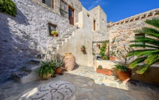 Stone residence facades and an exterior stairway surrounding a planted courtyard with terracotta pots and a circular pebble mosaic at Castle Residence in Monemvasia.