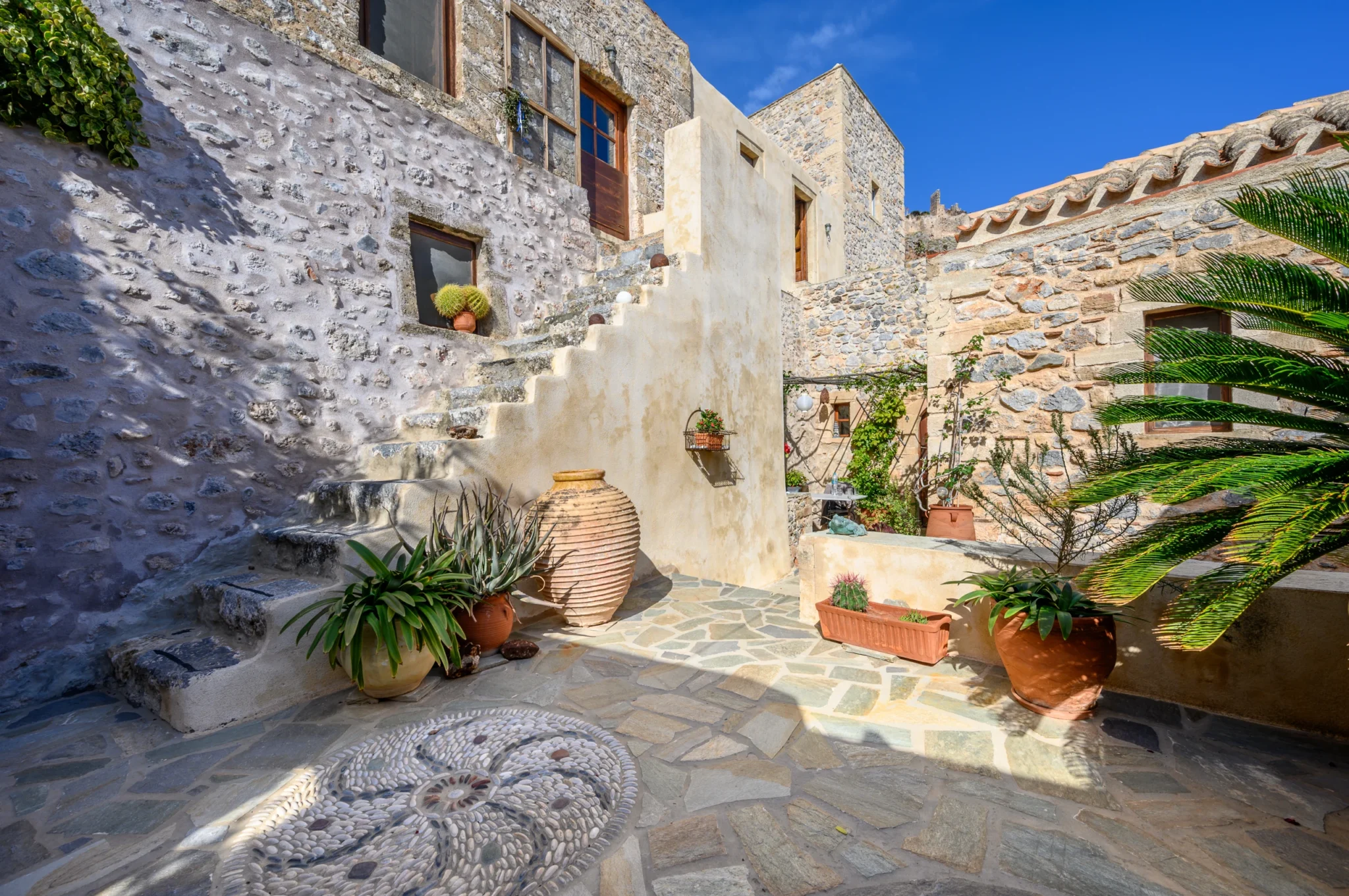 Stone residence facades and an exterior stairway surrounding a planted courtyard with terracotta pots and a circular pebble mosaic at Castle Residence in Monemvasia.