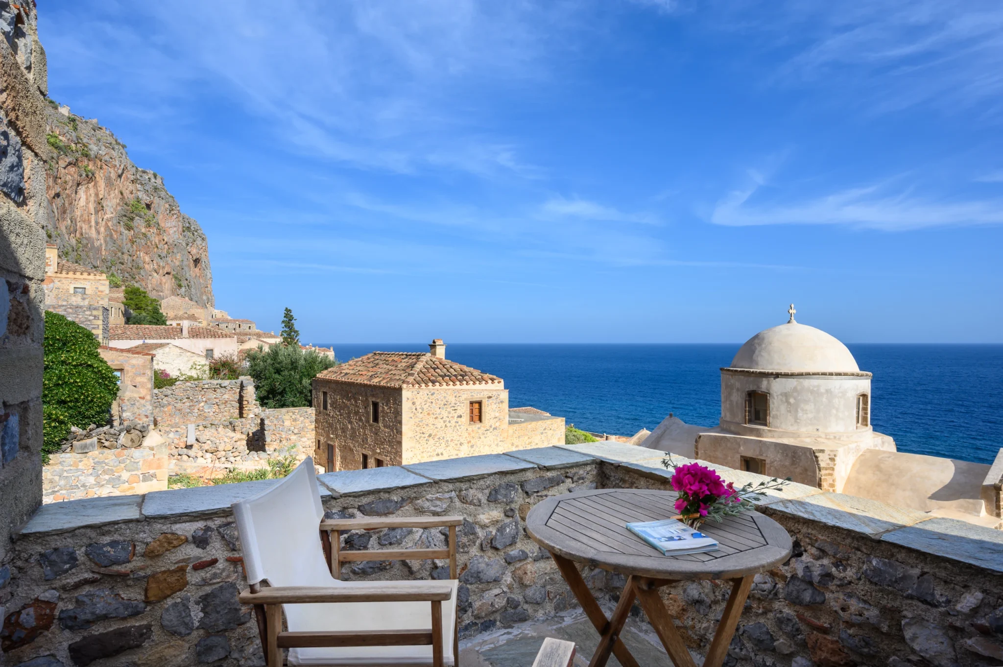 Round timber table with flowers and a canvas chair on a stone terrace overlooking Monemvasia rooftops, a church dome and the sea at Castle Residence in Monemvasia.