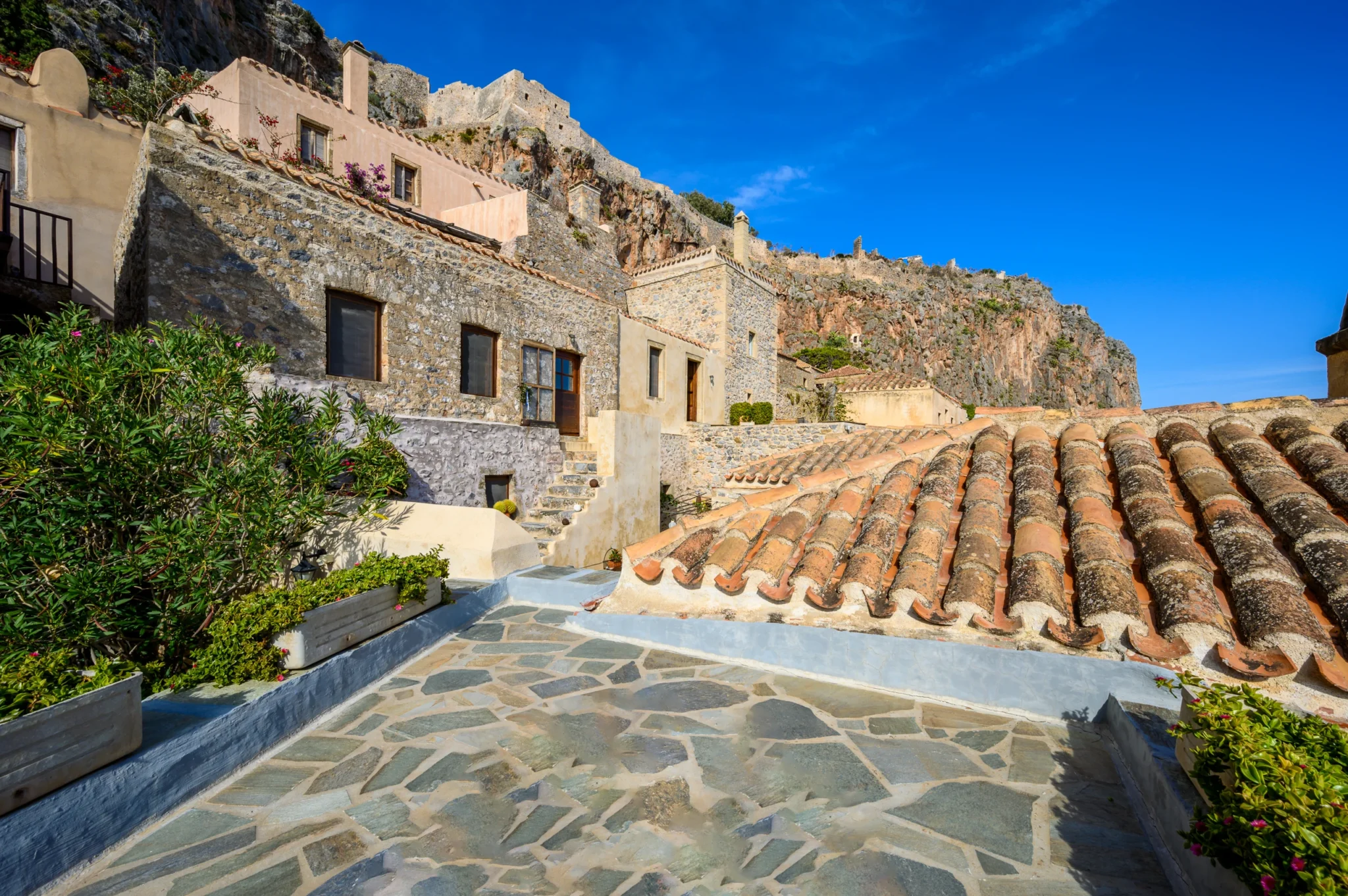 Stone-paved terrace with planting and terracotta rooftops beneath Monemvasia rock and fortress walls at Castle Residence in Monemvasia.