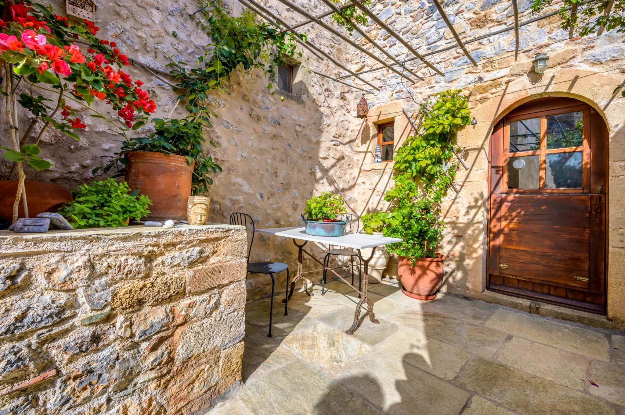 Small courtyard with a bistro table, climbing greenery, bougainvillea and an arched timber door beneath a trellis at Castle Residence in Monemvasia.