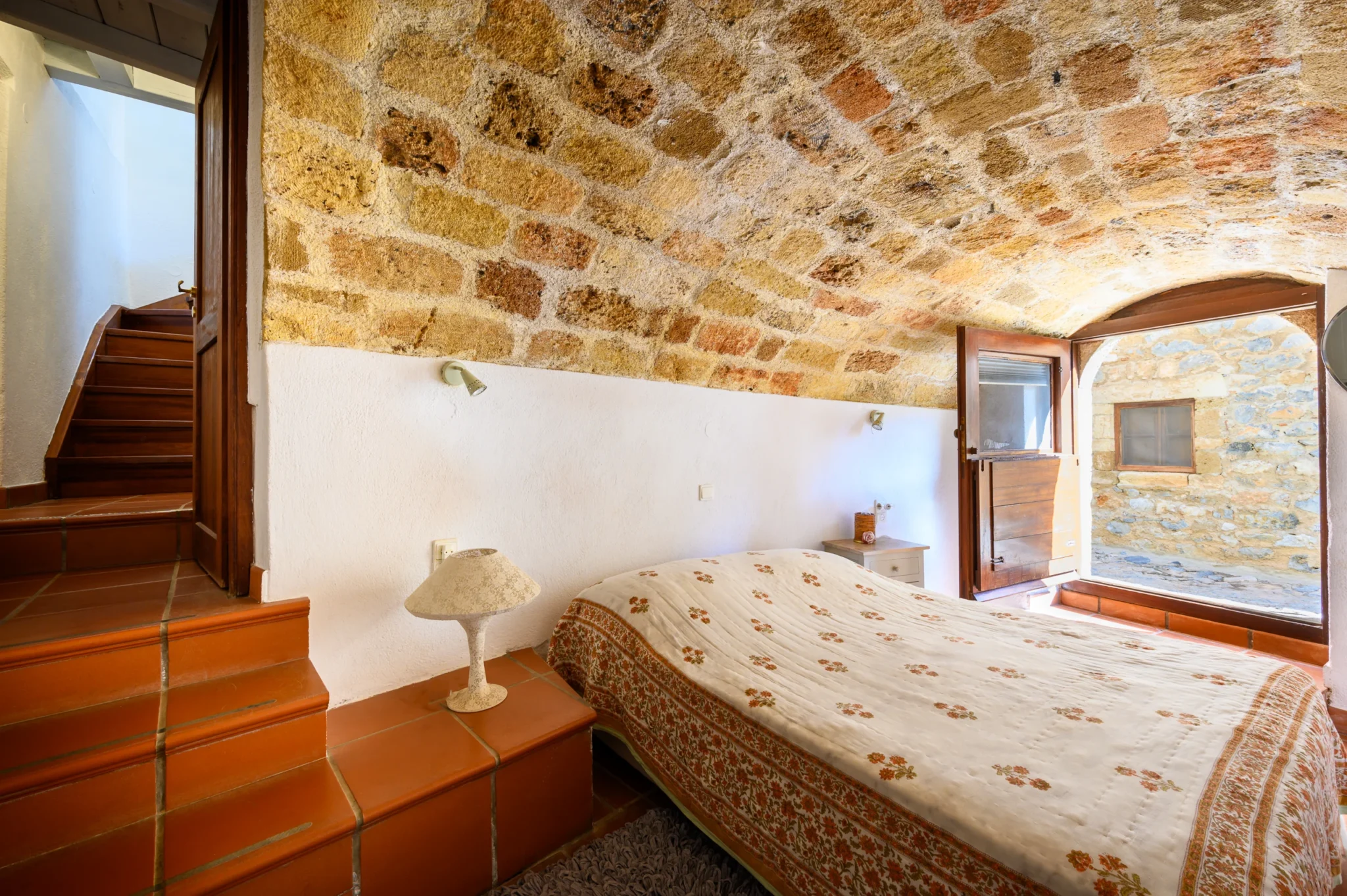 Bedroom beneath an exposed stone vault with a floral bedspread, terracotta steps and an open arched timber doorway at Castle Residence in Monemvasia.