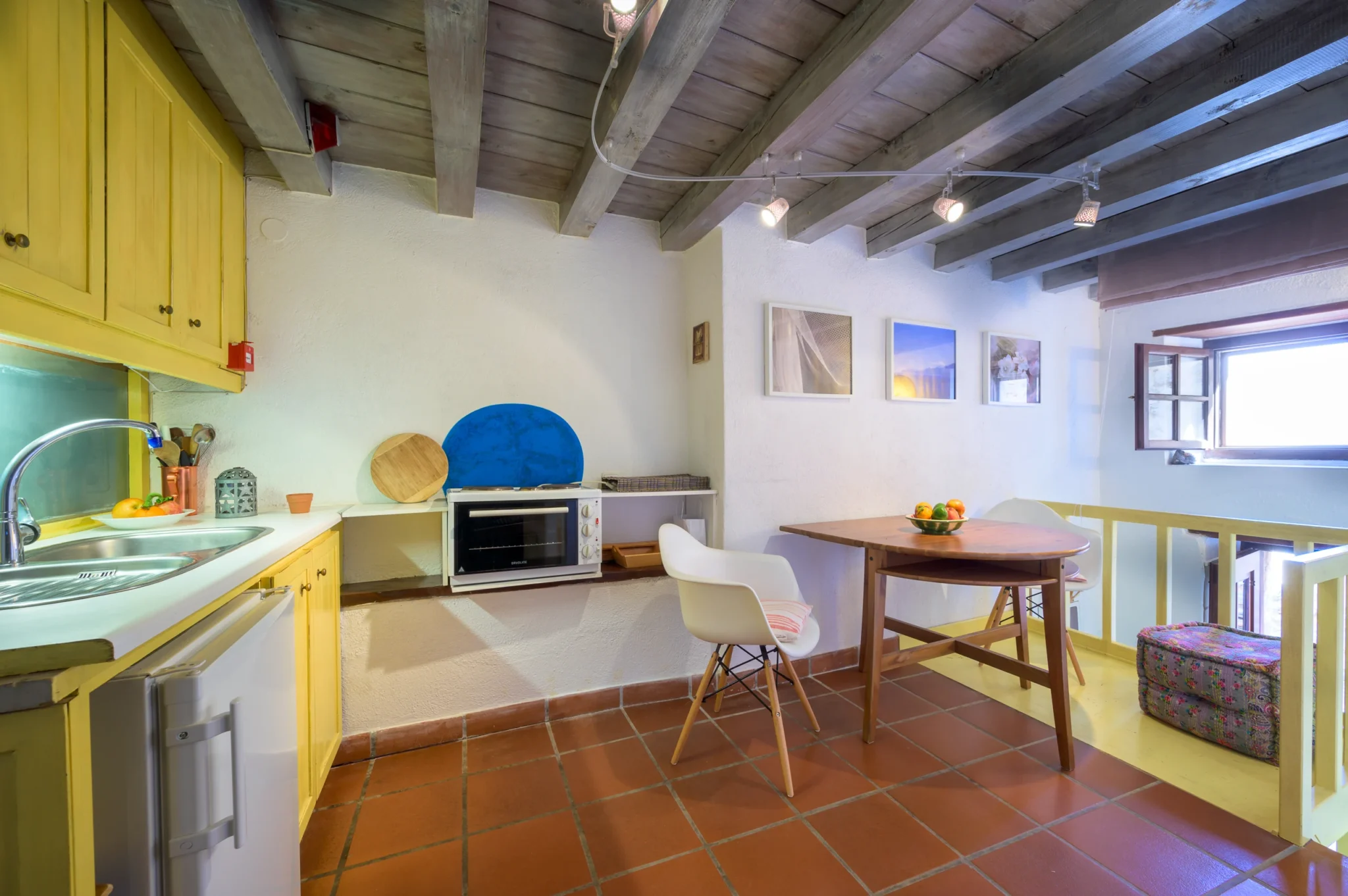Yellow kitchen cabinets and a timber dining table with white chairs beneath pale ceiling beams, beside a stairwell at Castle Residence in Monemvasia.