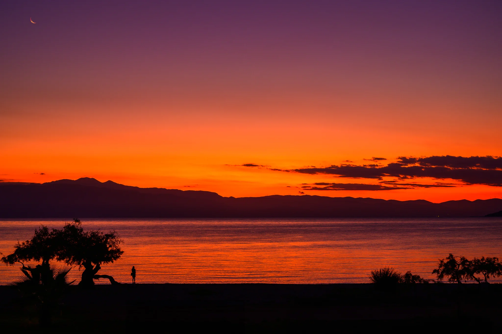 Crescent moon and lone figure beneath an intense red-orange coastal sunset