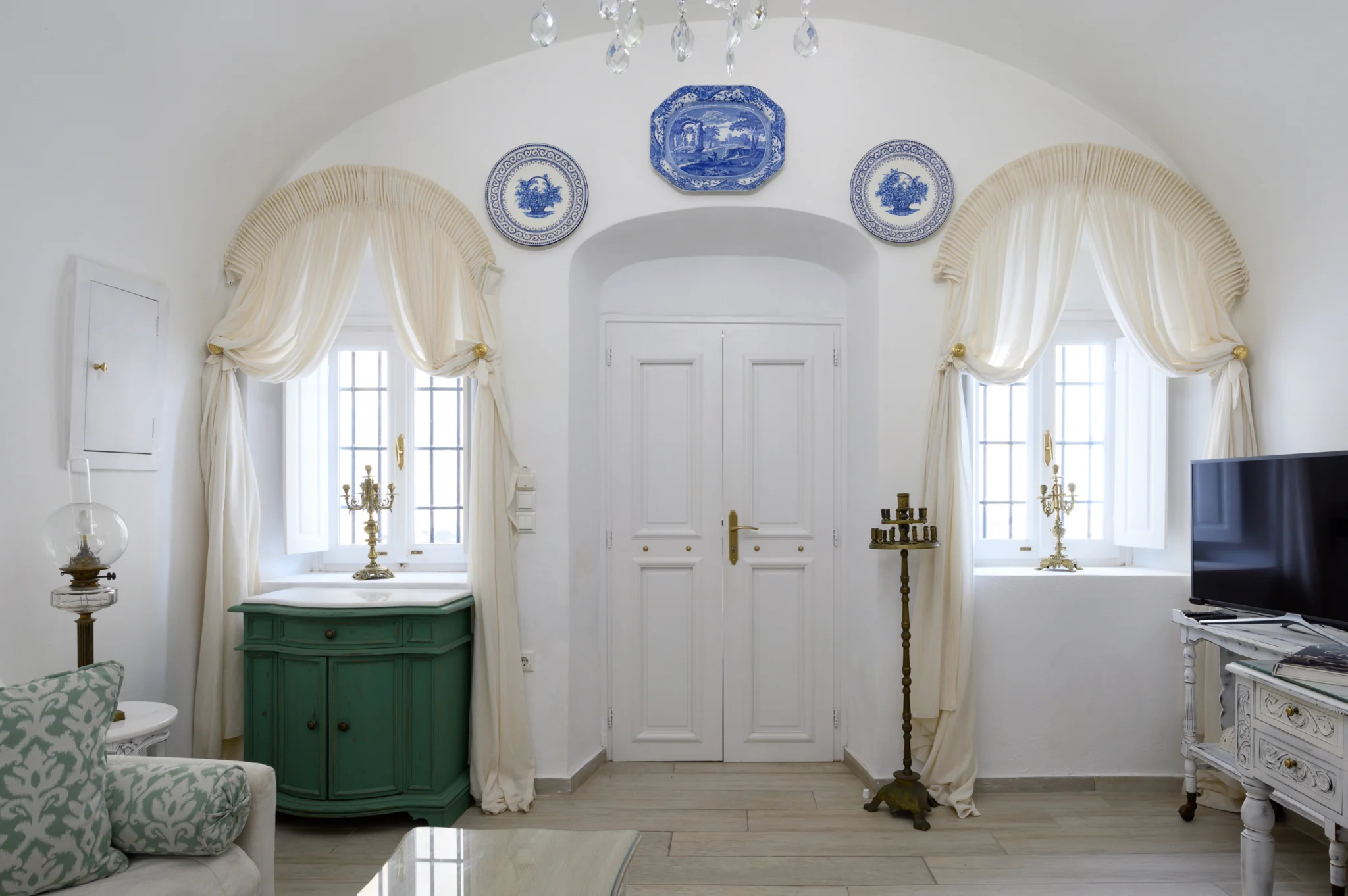 Intermezzo, Santorini — White entrance doors framed by draped windows, decorative plates and a green cabinet White entrance doors framed by draped windows, decorative plates and a green cabinet at Intermezzo in Santorini.