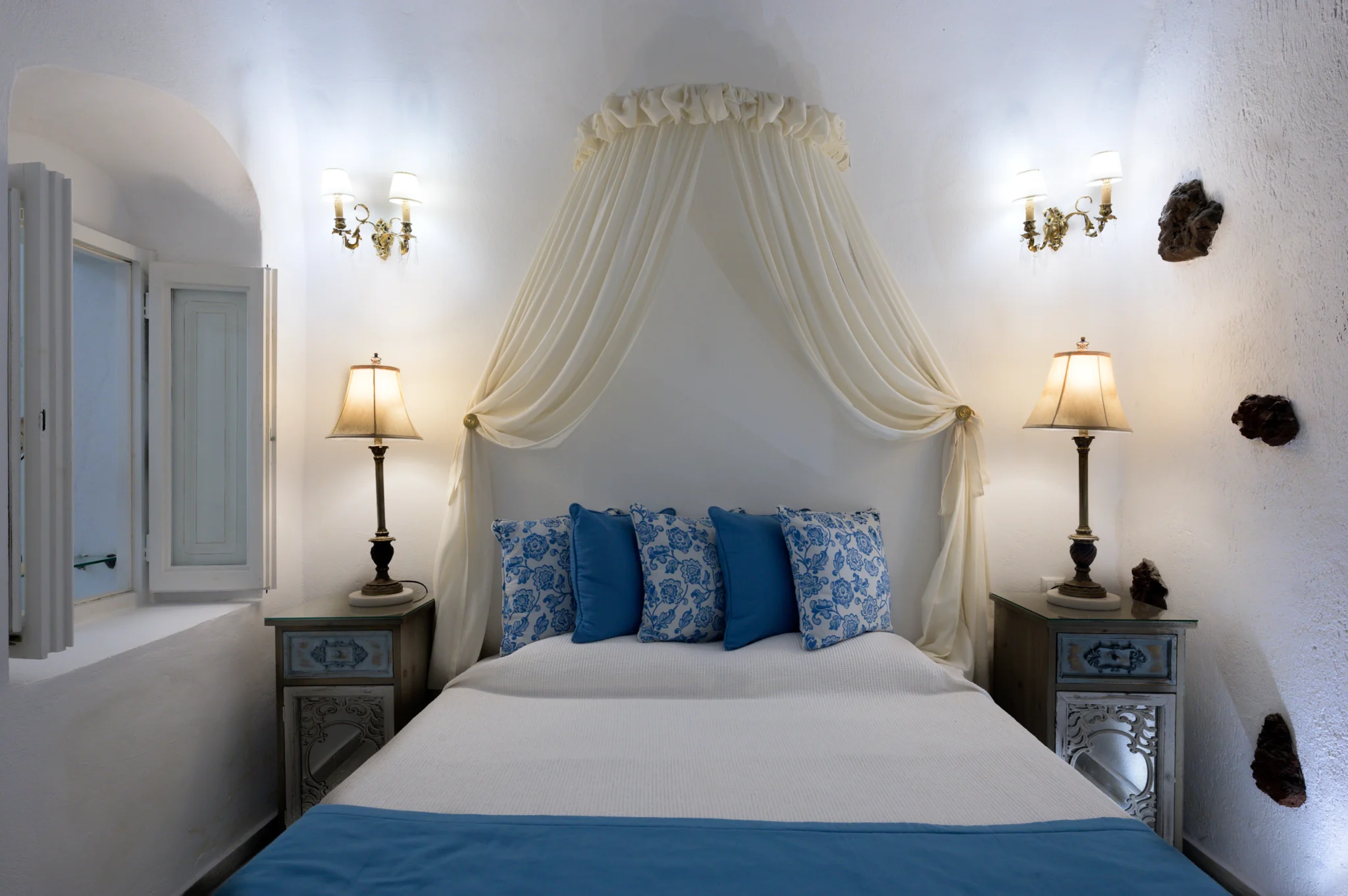 Intermezzo, Santorini — Bedroom with blue cushions, a draped canopy and paired bedside lamps Bedroom with blue cushions, a draped canopy and paired bedside lamps at Intermezzo in Santorini.