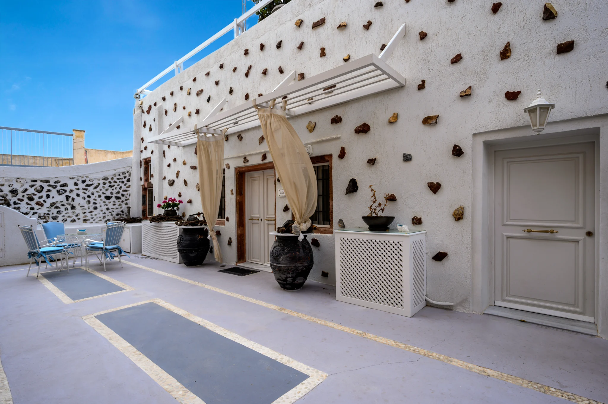 Intermezzo, Santorini — Courtyard and white facade with stone inserts, pergola curtains and outdoor seating Courtyard and white facade with stone inserts, pergola curtains and outdoor seating at Intermezzo in Santorini.