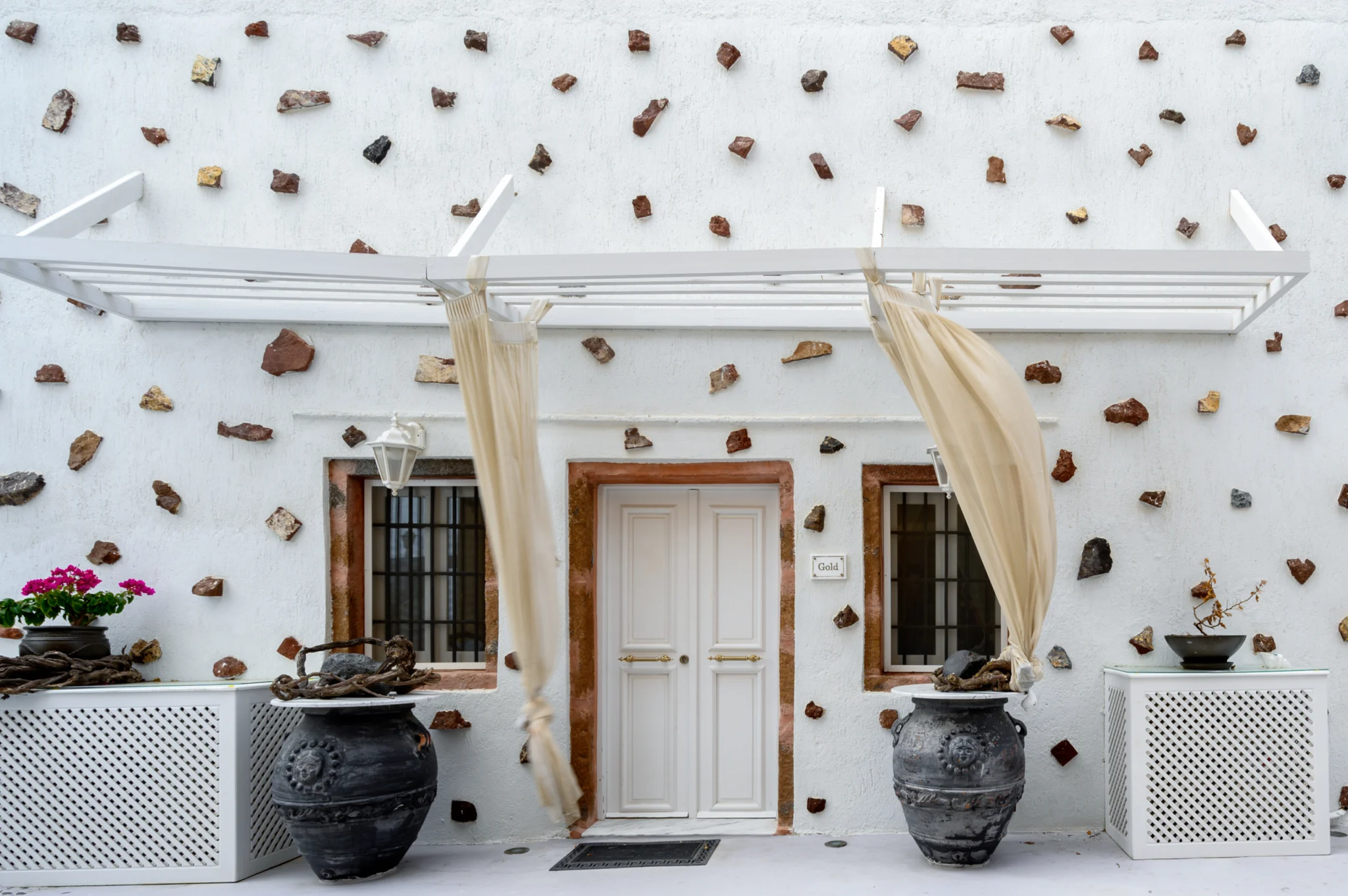 Front entrance with white pergola, flowing curtains and two large dark decorative pots at Intermezzo in Santorini.