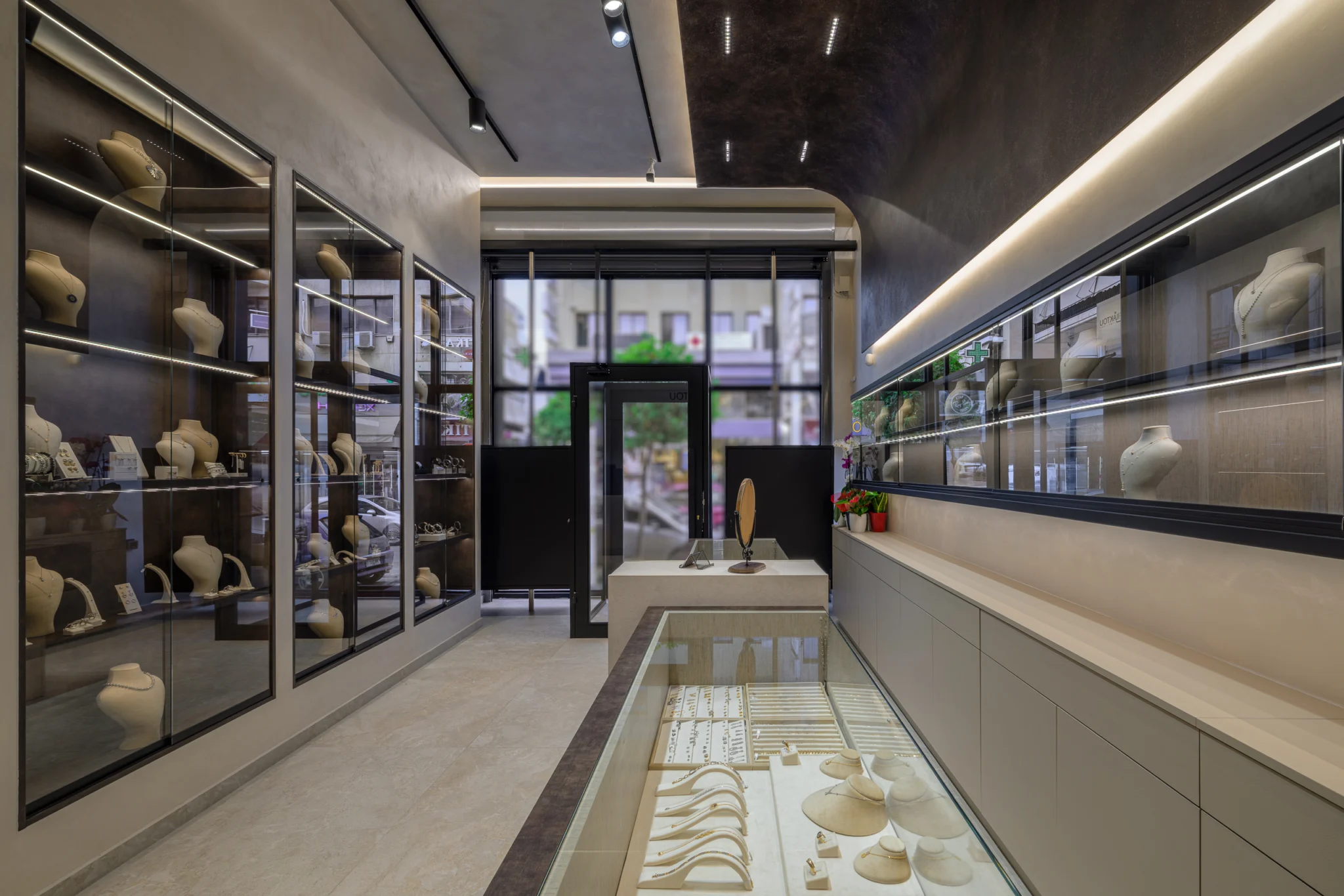 View towards the glazed entrance of a Korydallos jewelry shop, between illuminated wall cabinets and a central jewelry display.