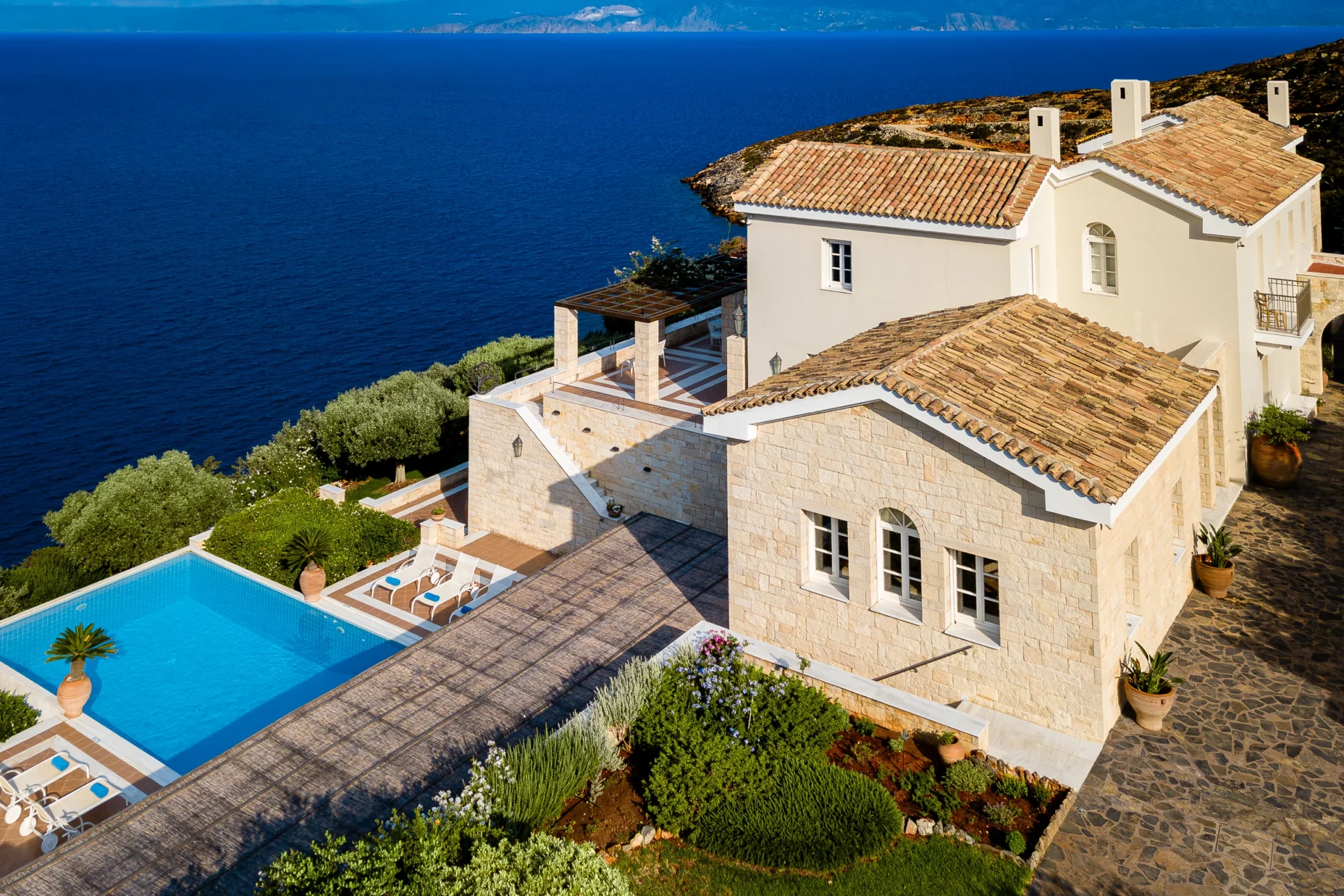 Aerial view of Mansion Olous in Elounda, showing tiled roofs, stone facades, planted terraces and a swimming pool beside the sea.
