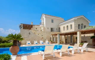 Stone and cream-coloured facade of Mansion Olous above a blue swimming pool, with timber pergolas, white sun loungers and terracotta roofs in Elounda.