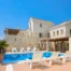 Stone and cream-coloured facade of Mansion Olous above a blue swimming pool, with timber pergolas, white sun loungers and terracotta roofs in Elounda.