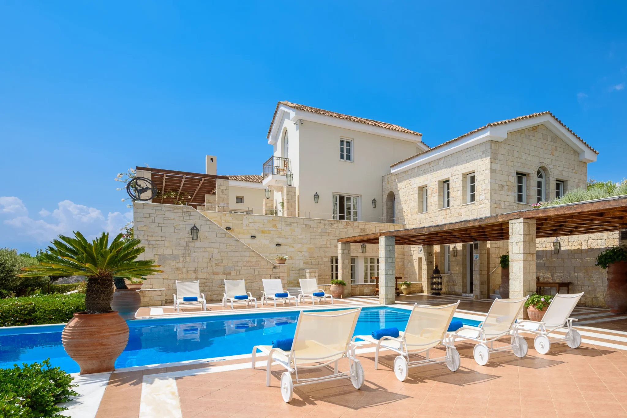 Stone and cream-coloured facade of Mansion Olous above a blue swimming pool, with timber pergolas, white sun loungers and terracotta roofs in Elounda.