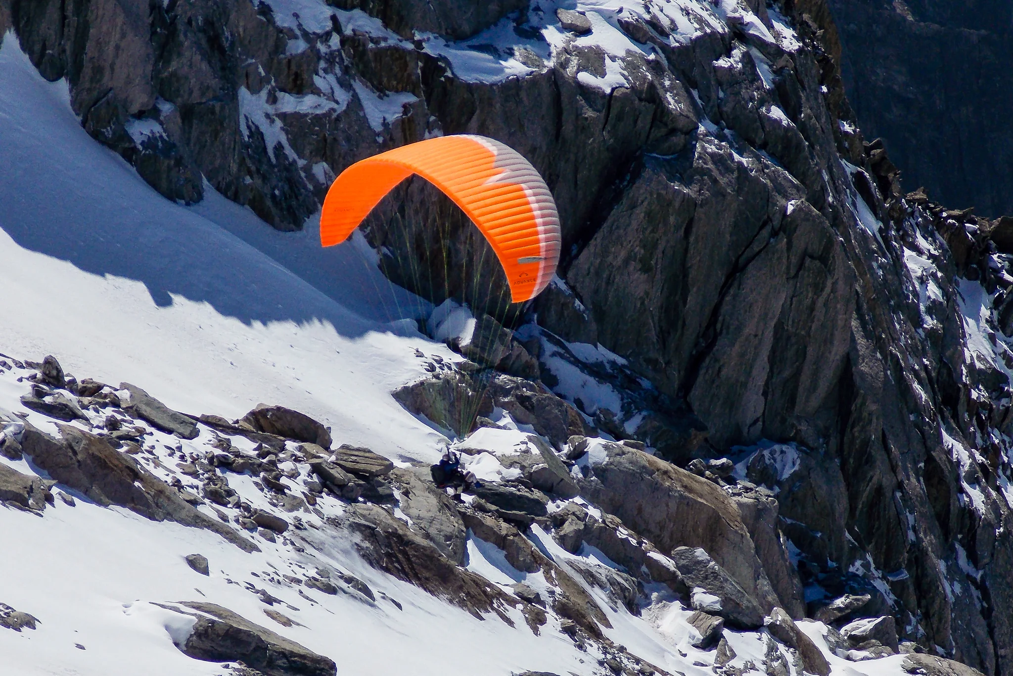 Orange paraglider crossing in front of snow-covered alpine rock