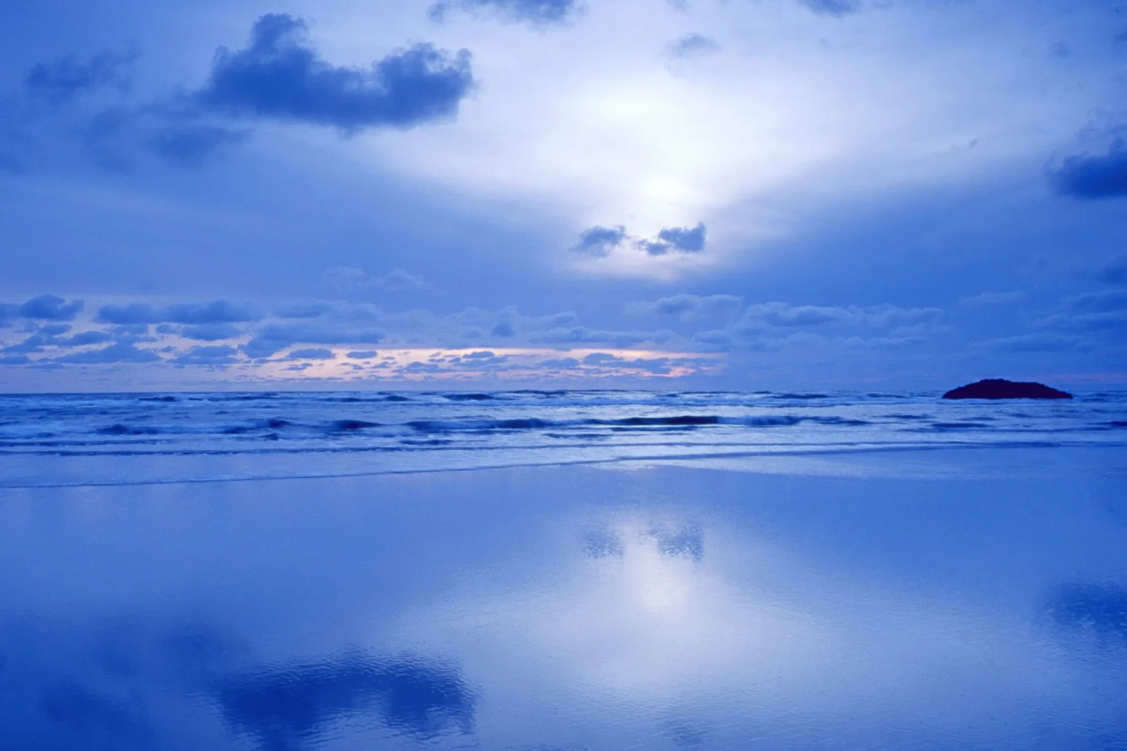 Reflective blue beach and small offshore rock beneath an overcast twilight sky