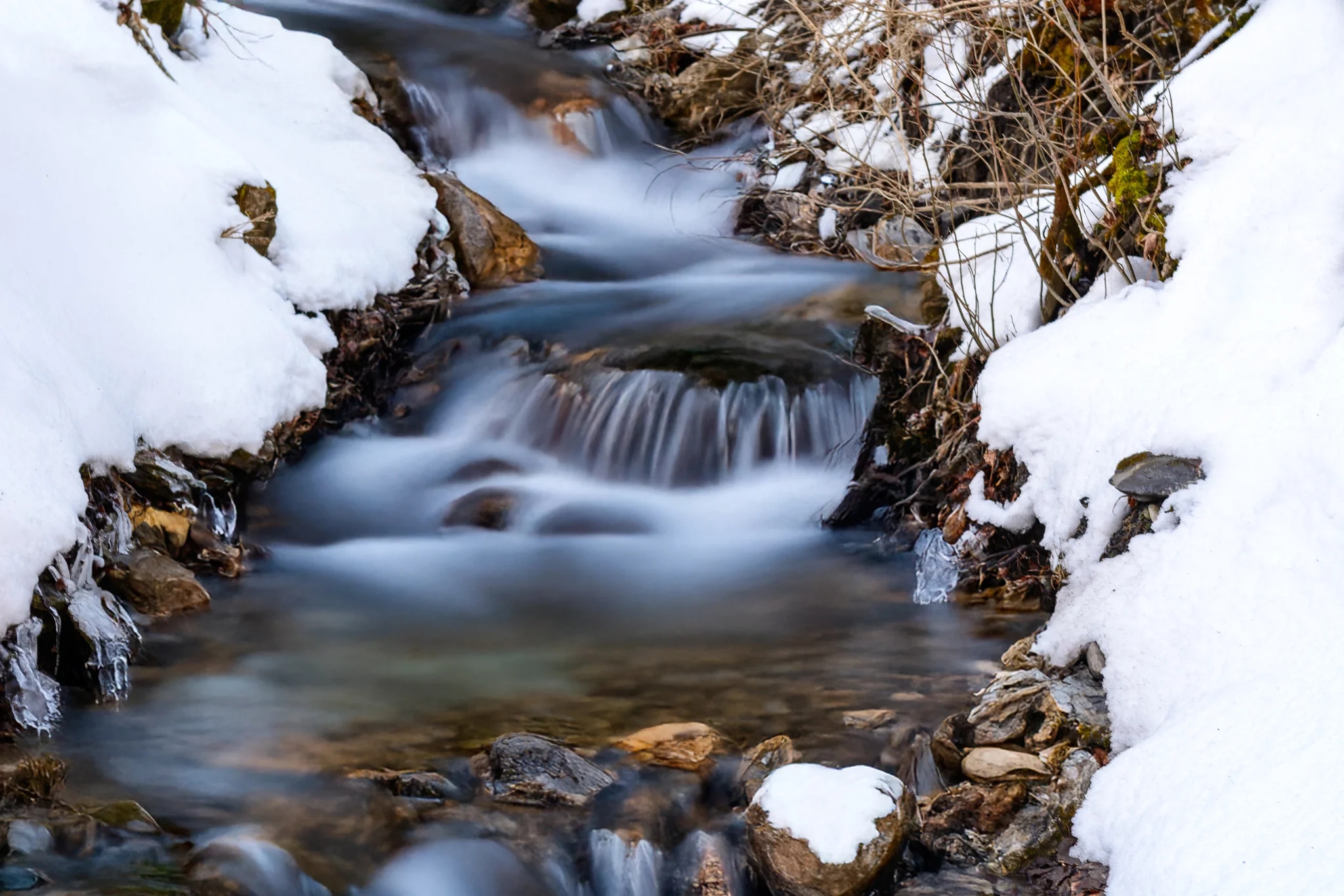 Long-exposure mountain stream flowing between banks of snow