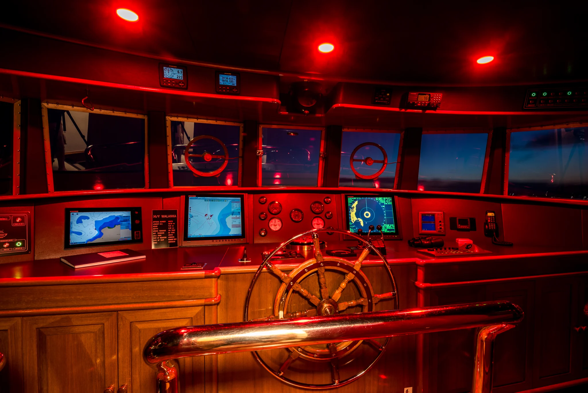 Yacht wheelhouse with navigation screens and a brass wheel under red lighting at night