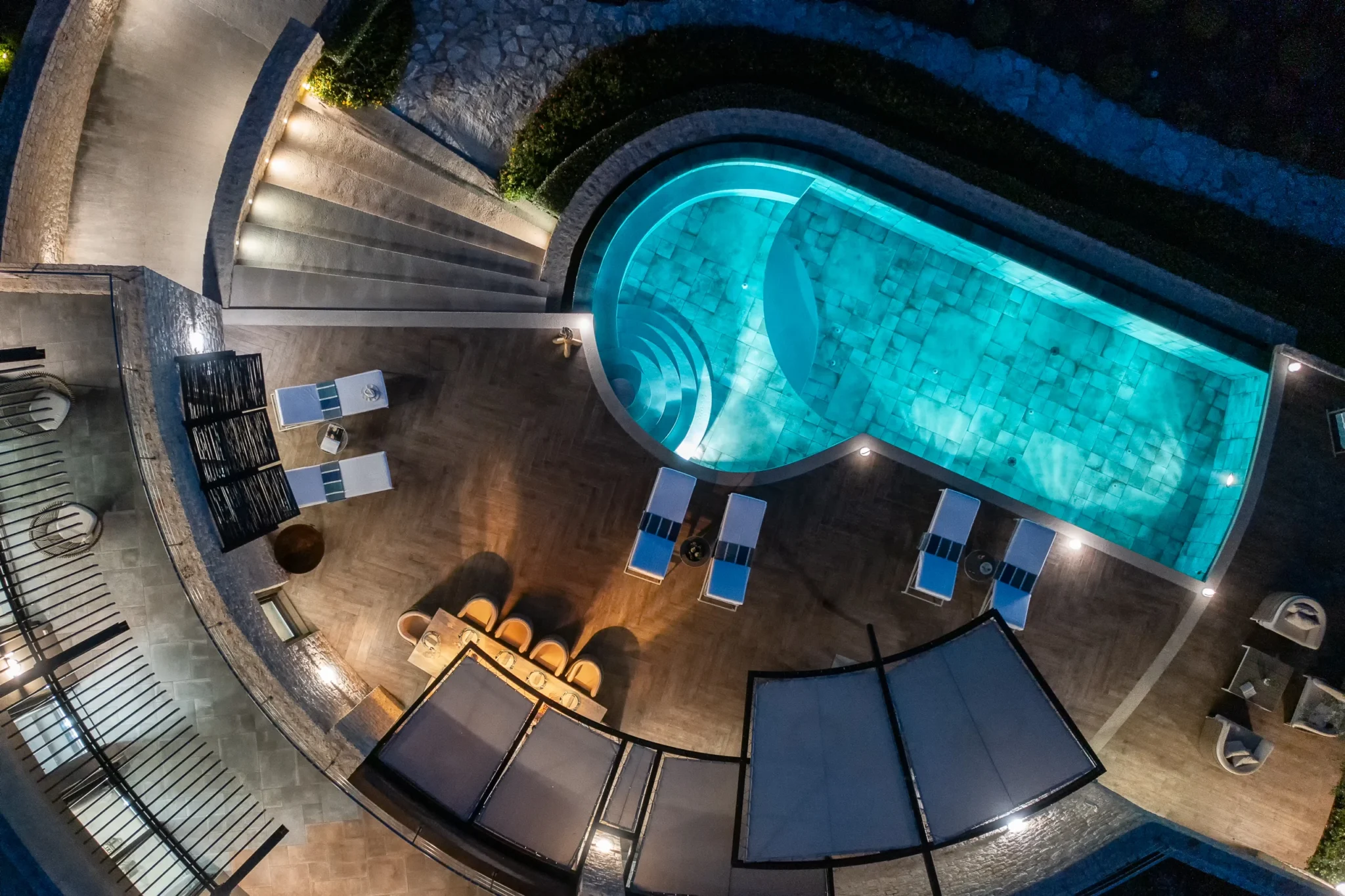 Overhead drone view of a turquoise illuminated swimming pool, curved steps, sun loungers and a covered dining terrace at night at The Olive Retreat near Pylos.