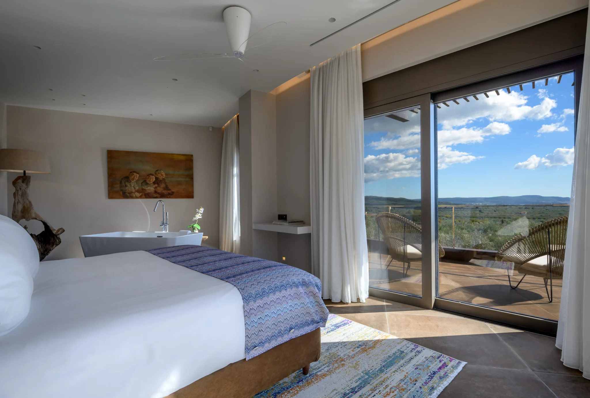 White bed with a purple patterned runner and a freestanding bath beside sliding glass doors framing terrace chairs and the landscape beyond at The Olive Retreat near Pylos.