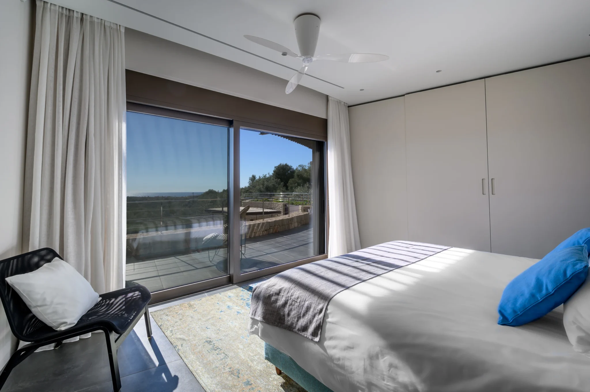 Bedroom with blue cushions, white wardrobes and a dark armchair beside sliding glass doors opening towards the terrace and olive trees at The Olive Retreat near Pylos.