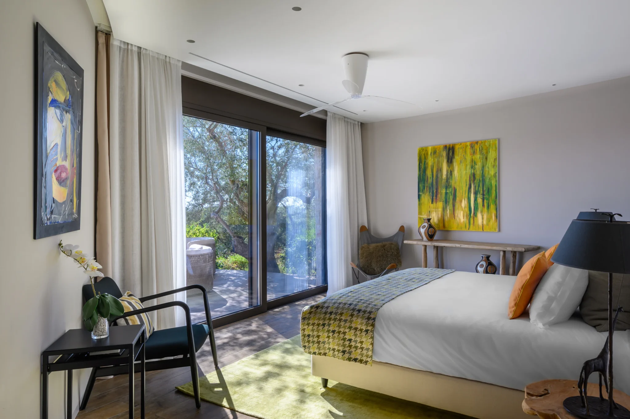 Bedroom with yellow cushions, a green patterned runner and abstract artwork, with a mature olive tree framed by tall glass doors at The Olive Retreat near Pylos.