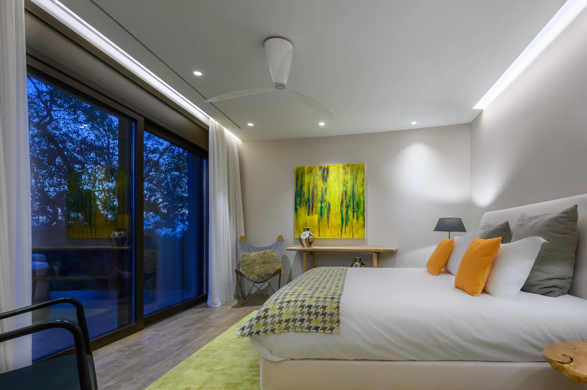 Illuminated bedroom with yellow cushions and abstract artwork, with olive trees silhouetted through the glass doors at blue hour at The Olive Retreat near Pylos.
