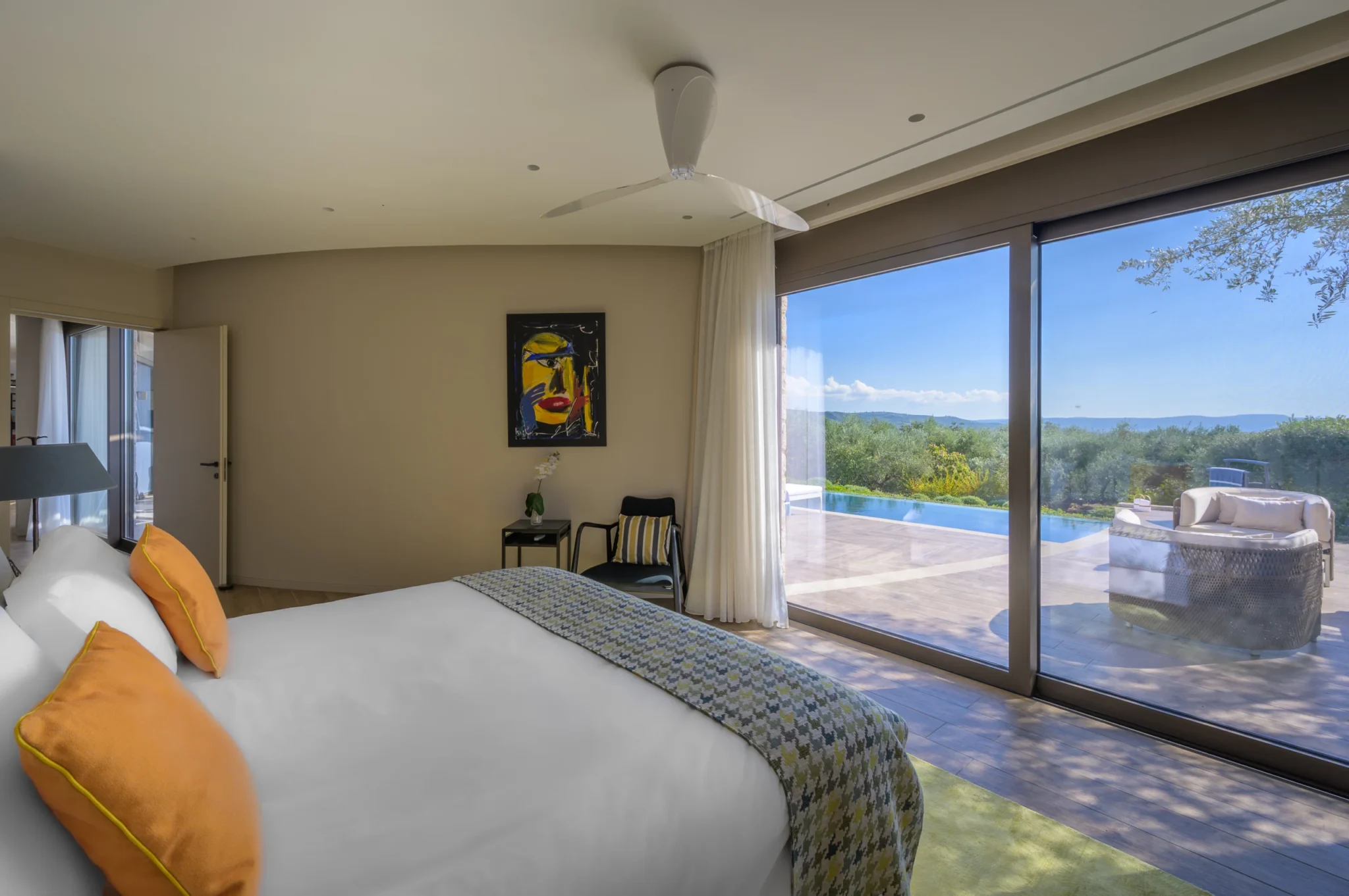 White bed with yellow cushions beside sliding glass doors overlooking the swimming pool, circular outdoor seating and olive trees at The Olive Retreat near Pylos.