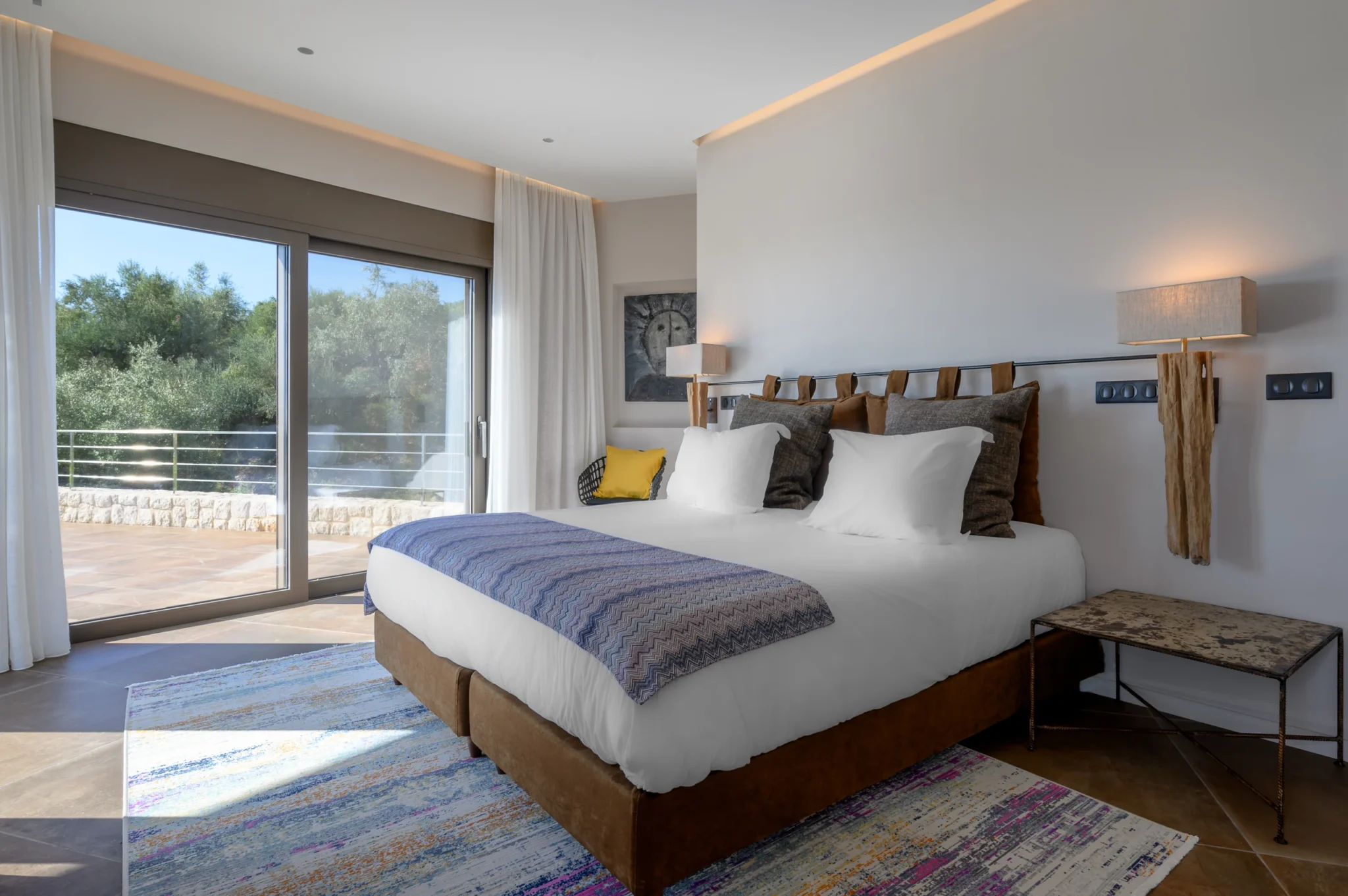 White bed with a purple patterned runner and timber wall lamps beside glass terrace doors overlooking olive trees at The Olive Retreat near Pylos.