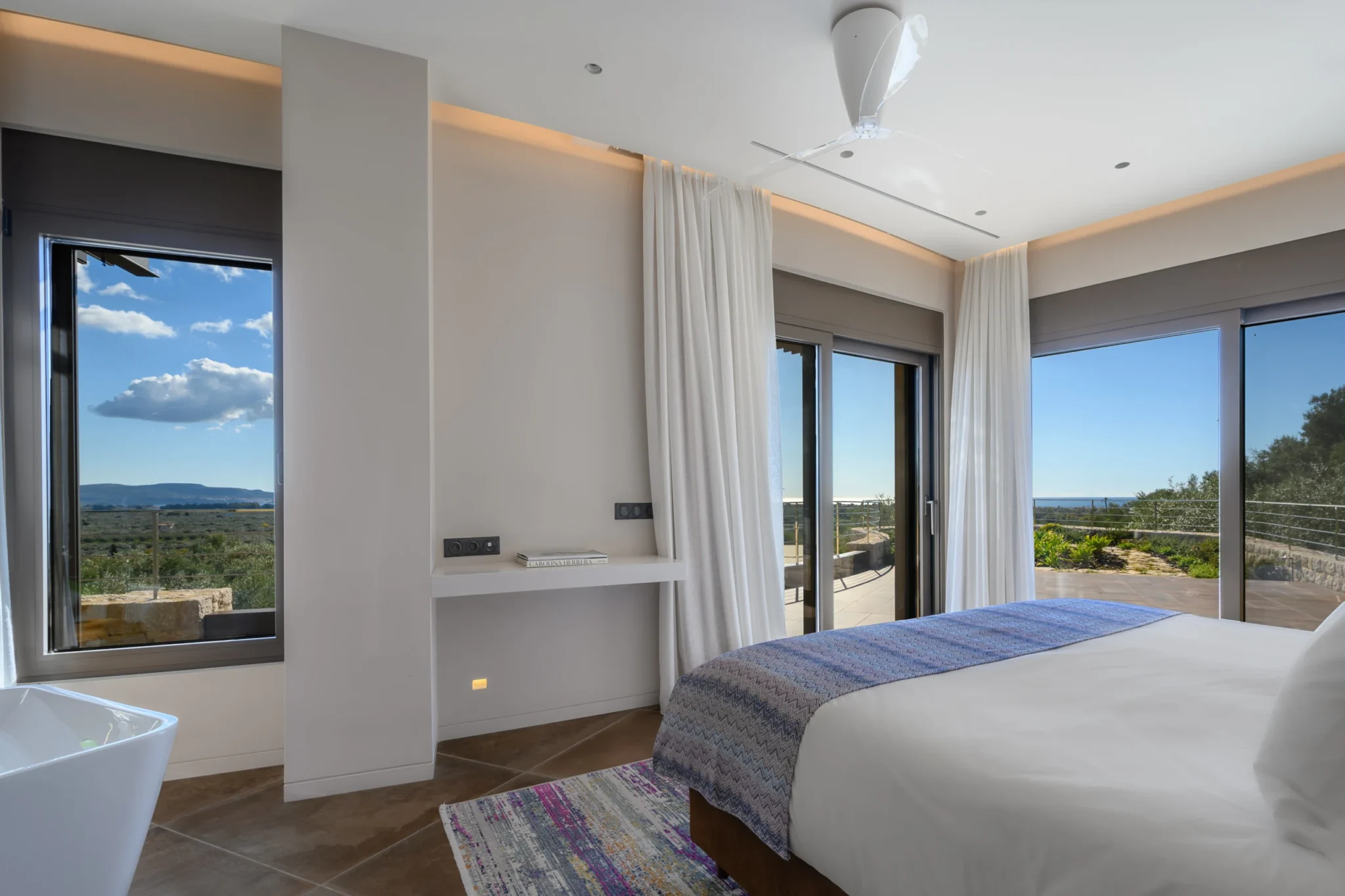White bed with a purple patterned runner beside tall windows and sliding doors overlooking the terrace and surrounding landscape at The Olive Retreat near Pylos.